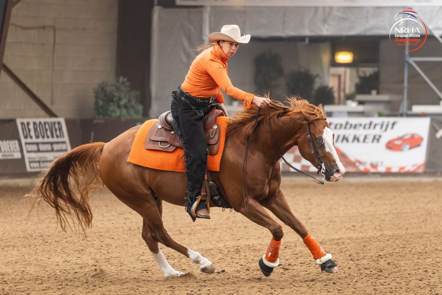 Rider in orange outfit performing reining on a chestnut horse in an indoor arena competition.