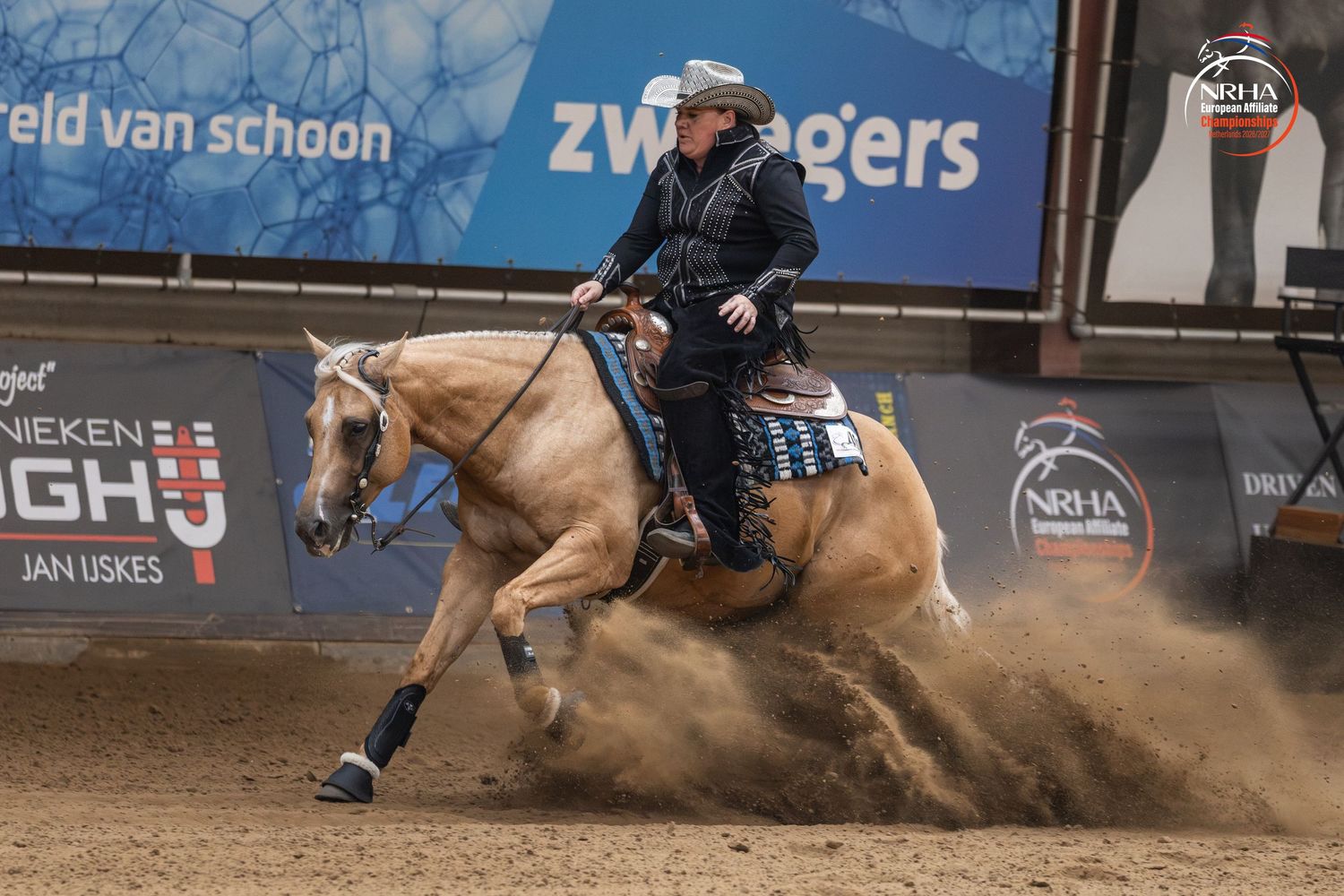 A rider on a palomino horse performs a sliding stop in an indoor reining competition with sponsors visible in background.