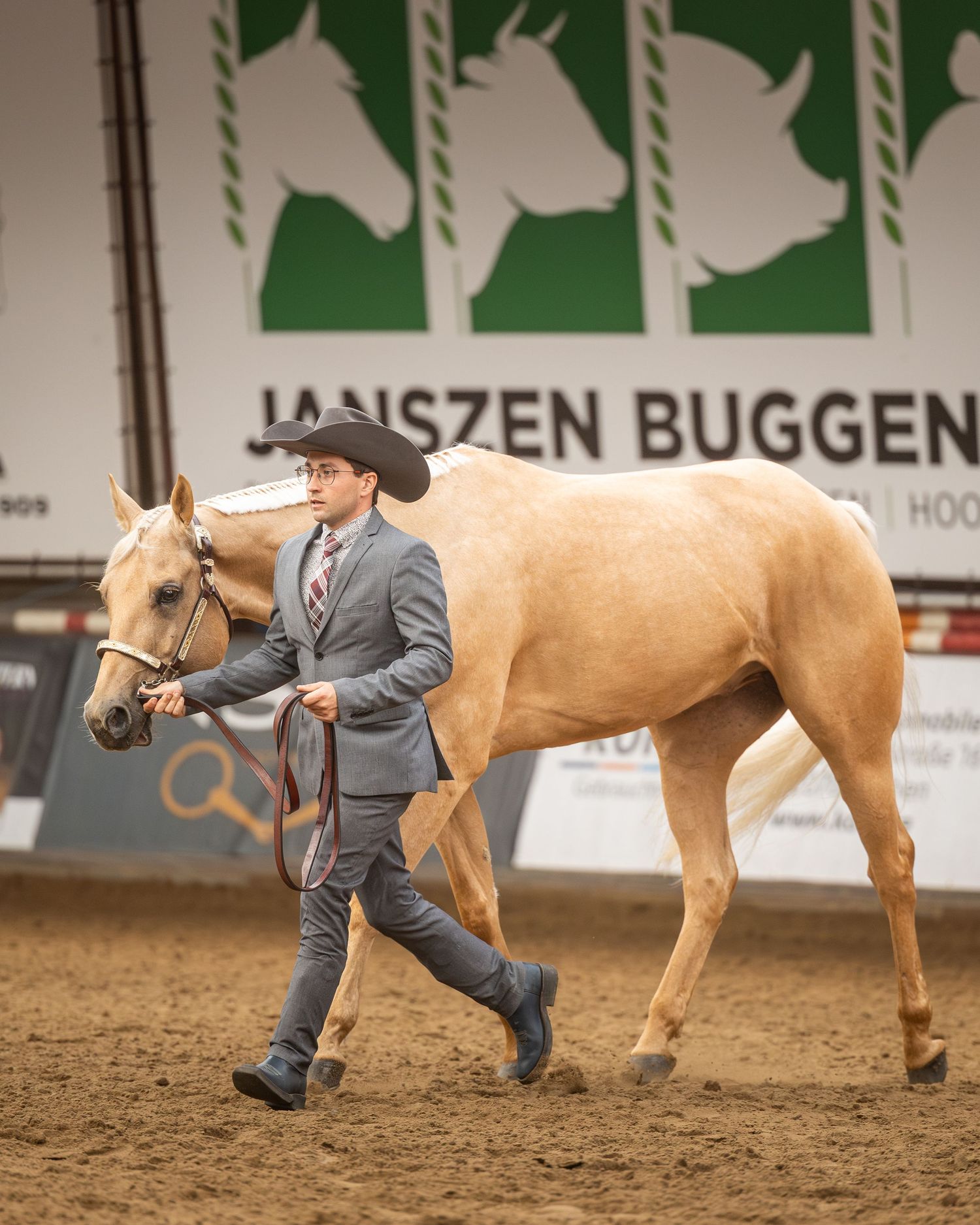 A young man in a gray suit and cowboy hat leads a palomino horse in an indoor equestrian show arena.