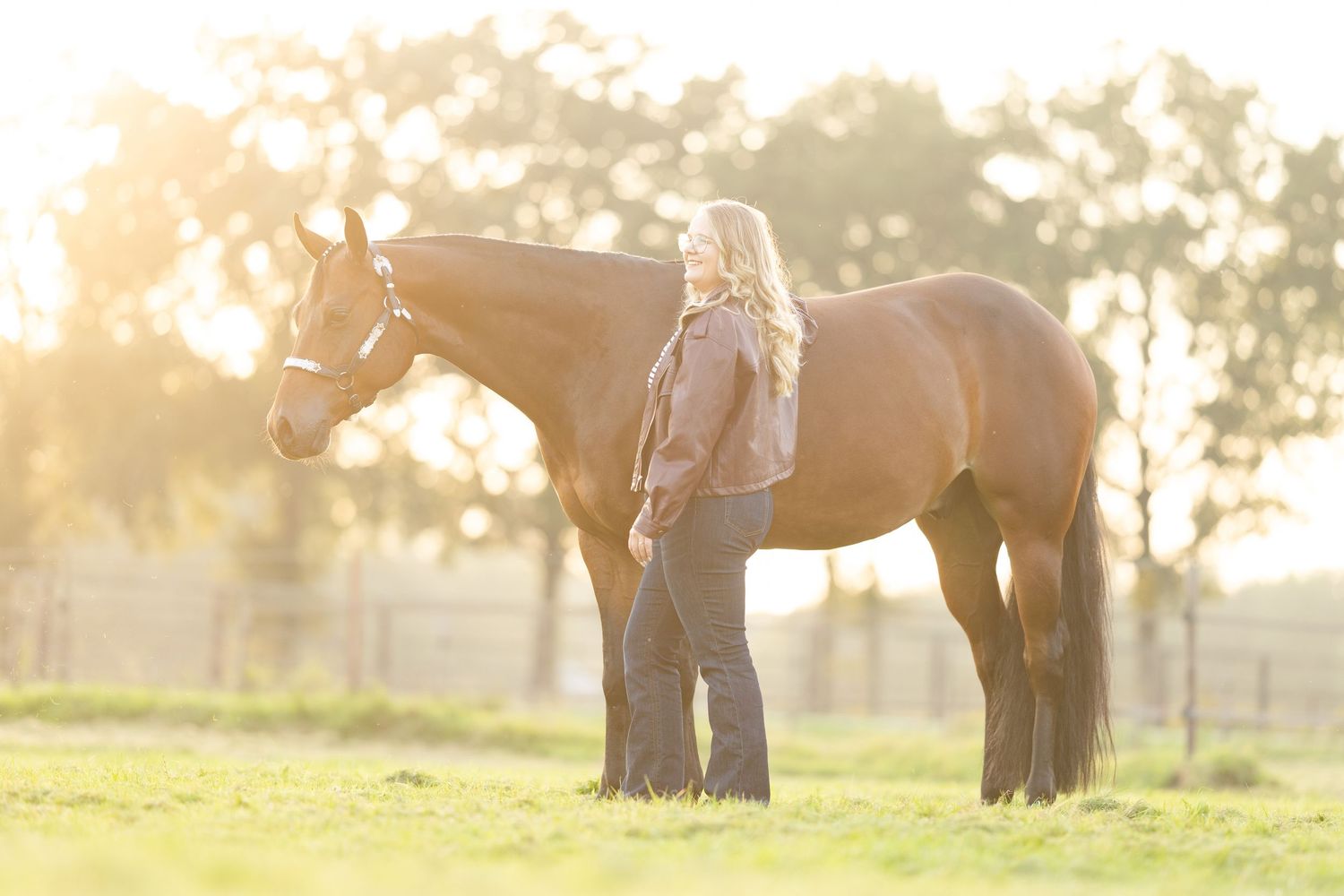 Woman standing beside a tall brown horse in a sunlit green pasture during golden hour.