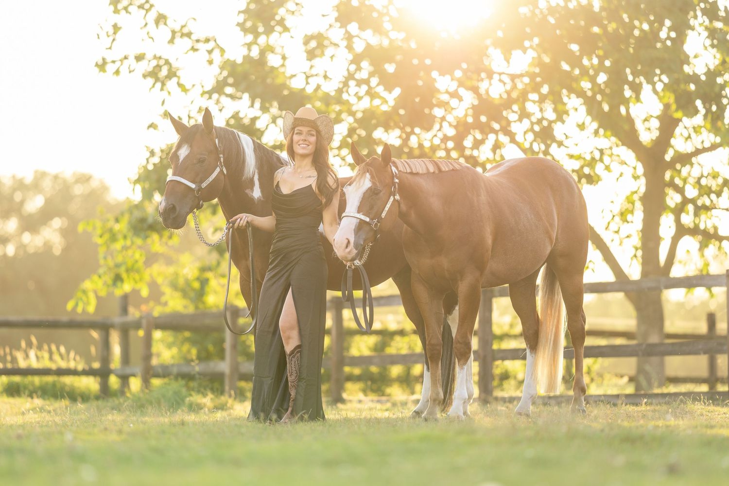 Woman in black gown poses with two horses at golden hour near a wooden fence in a sunlit field.