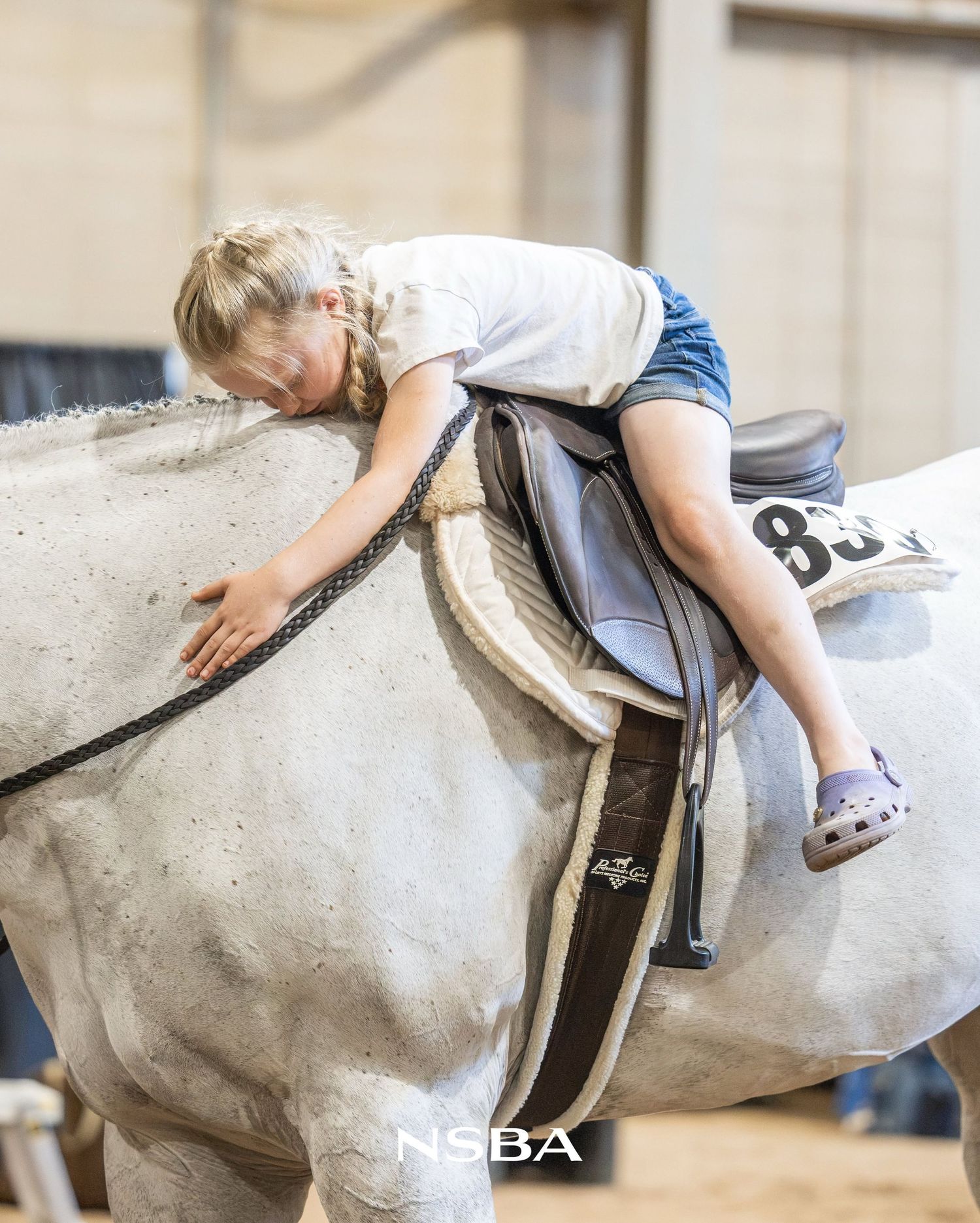 Young girl in denim shorts hugging a gray horse while seated in saddle at the NSBA World Show