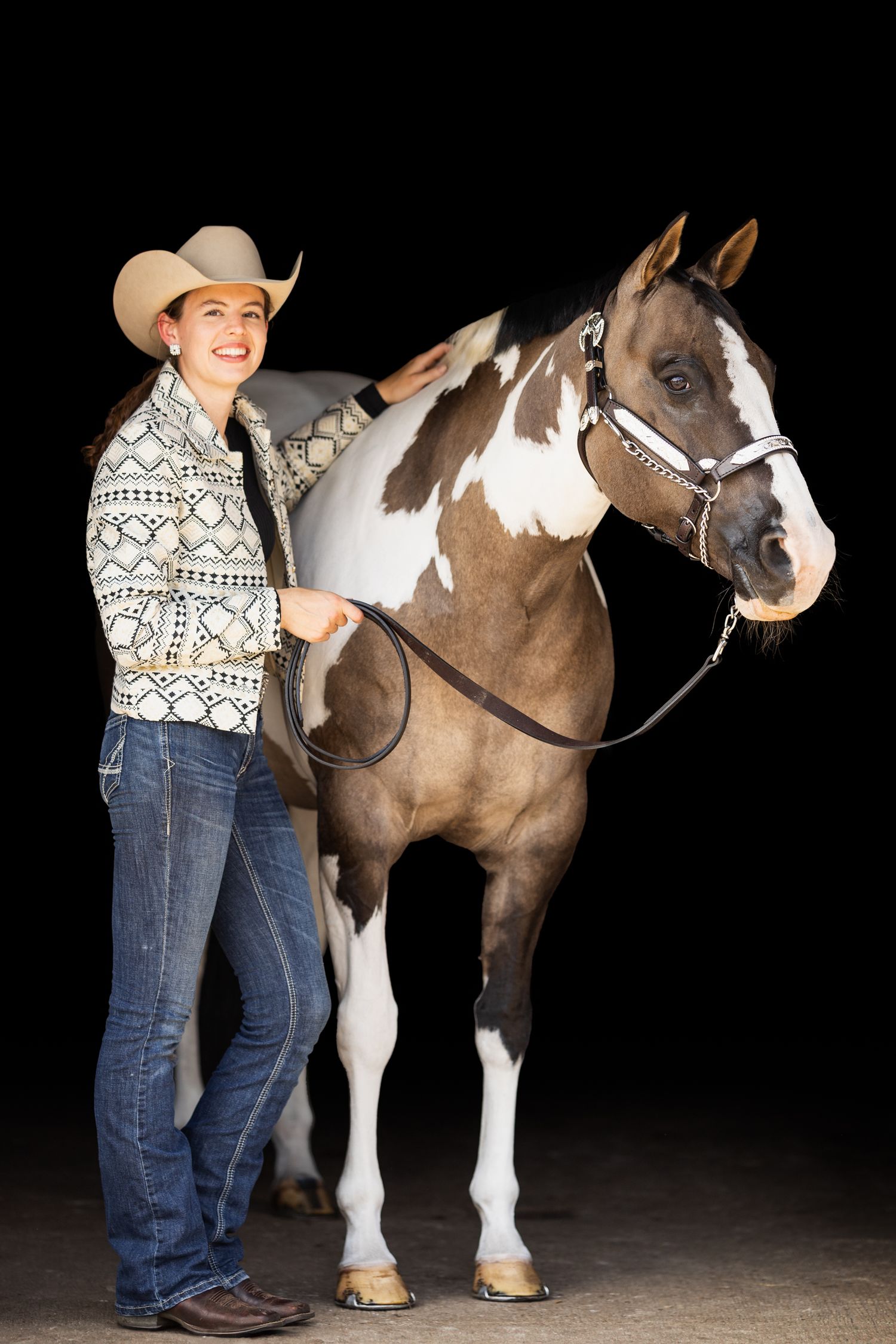 A paint horse poses with a rider in a patterned sweater and jeans against black backdrop.