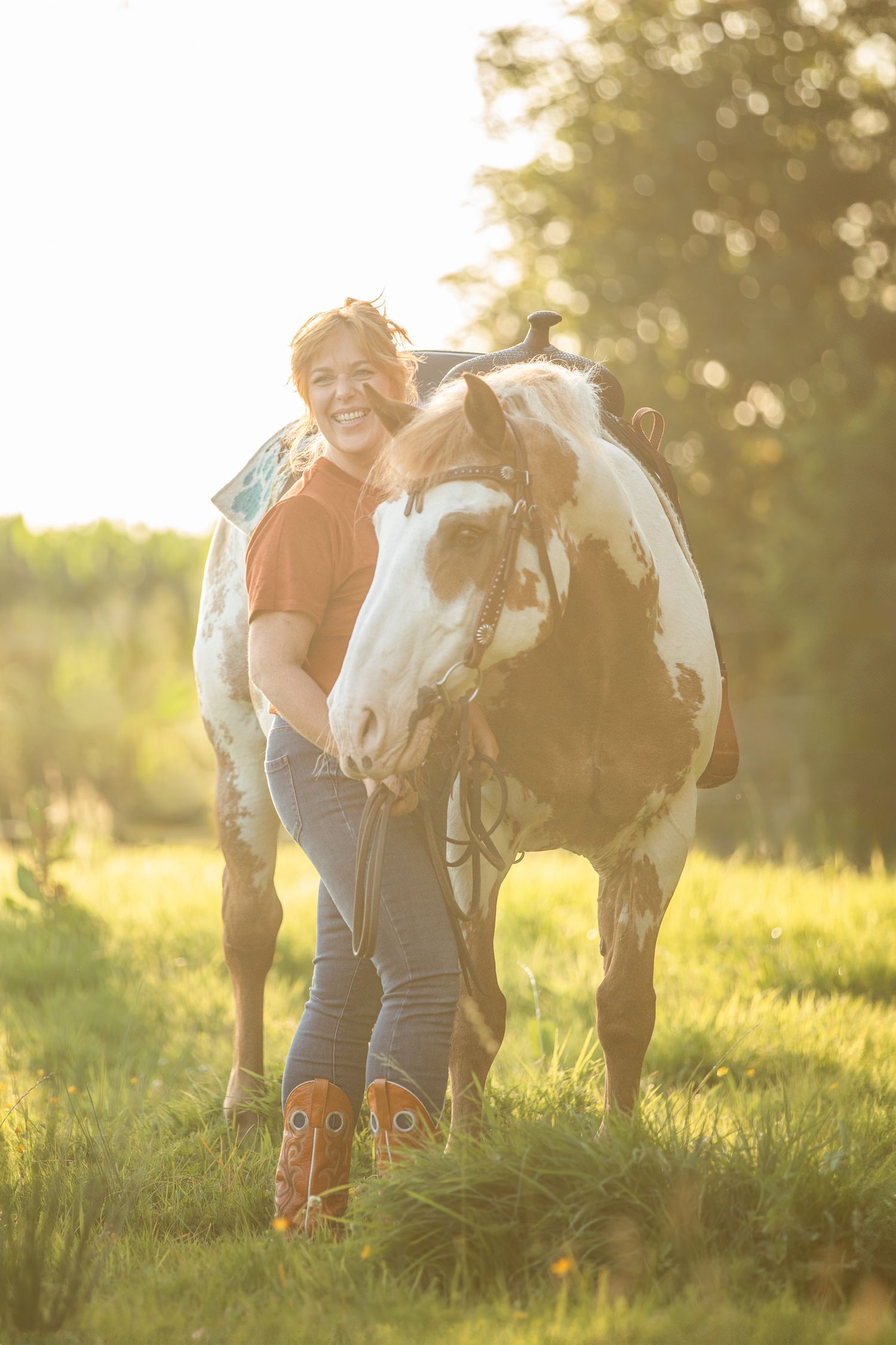 Person in orange shirt and jeans kneeling beside a paint horse in golden sunlit meadow.