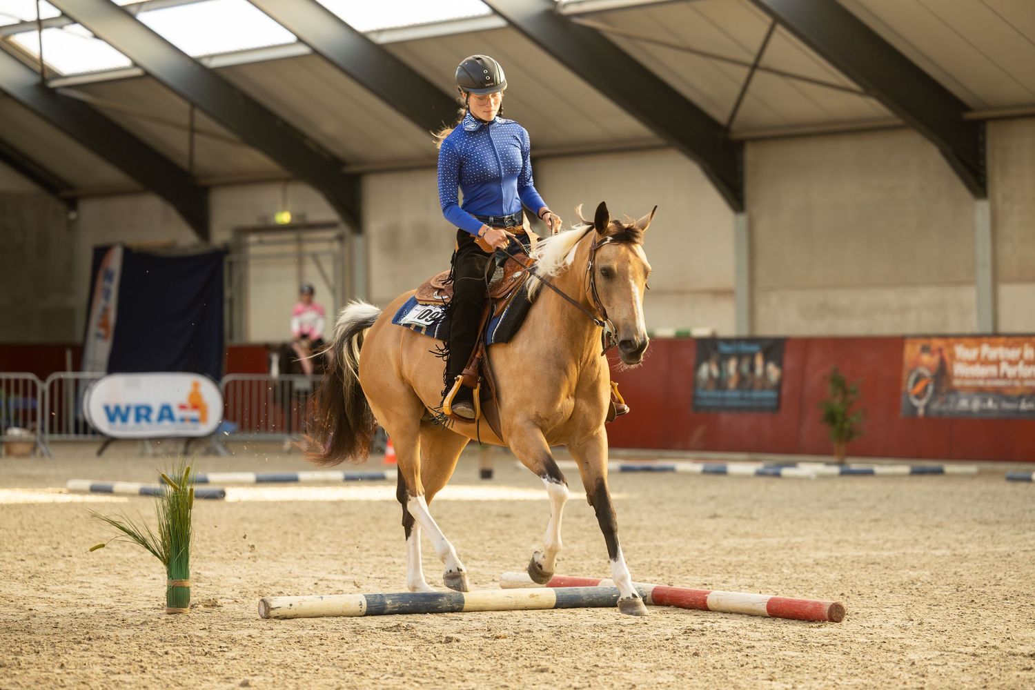 Multiple riders practicing jumps in an indoor arena with western riding equipment.