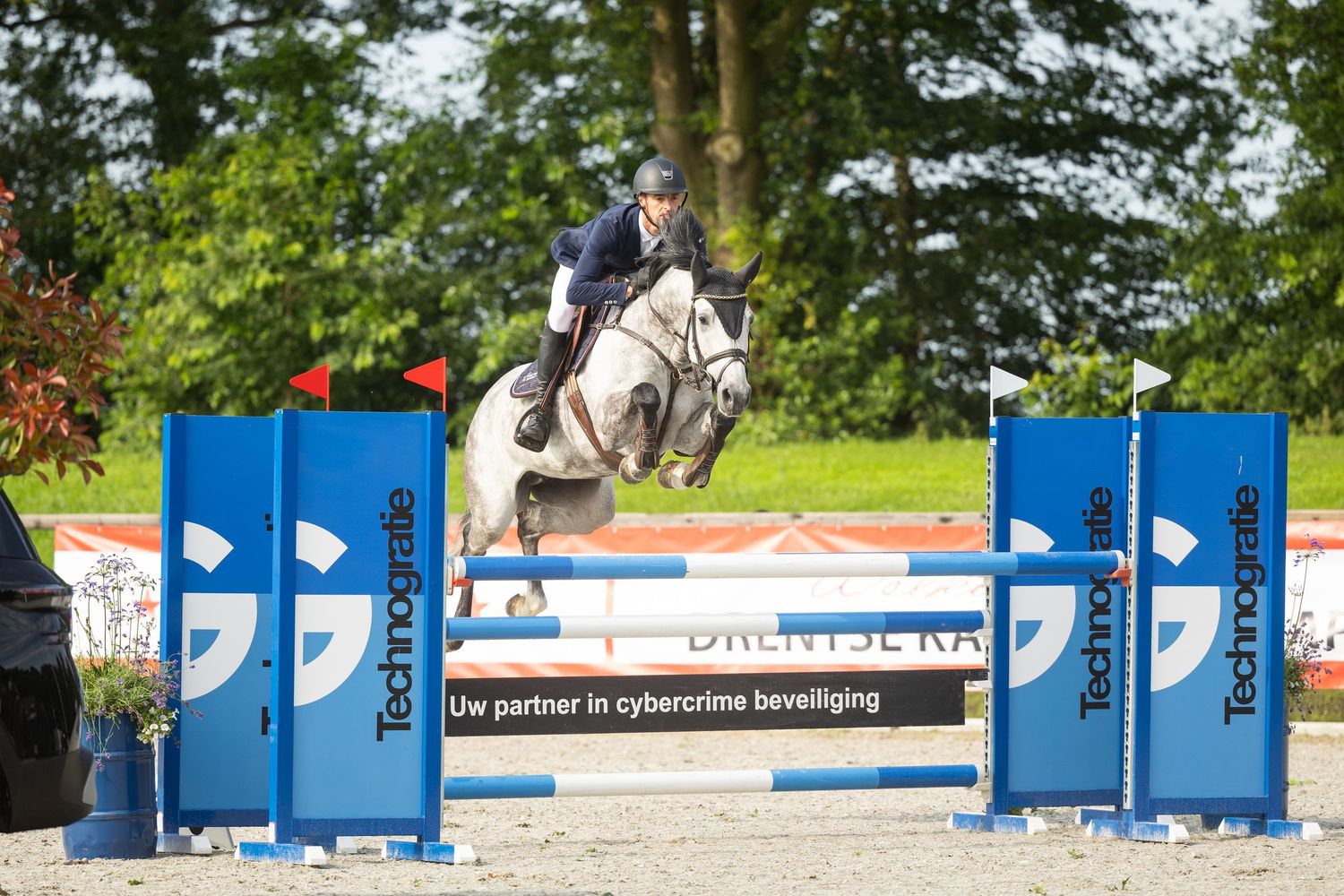 Equestrian rider jumping a gray horse over a colorful obstacle at an outdoor show jumping competition.