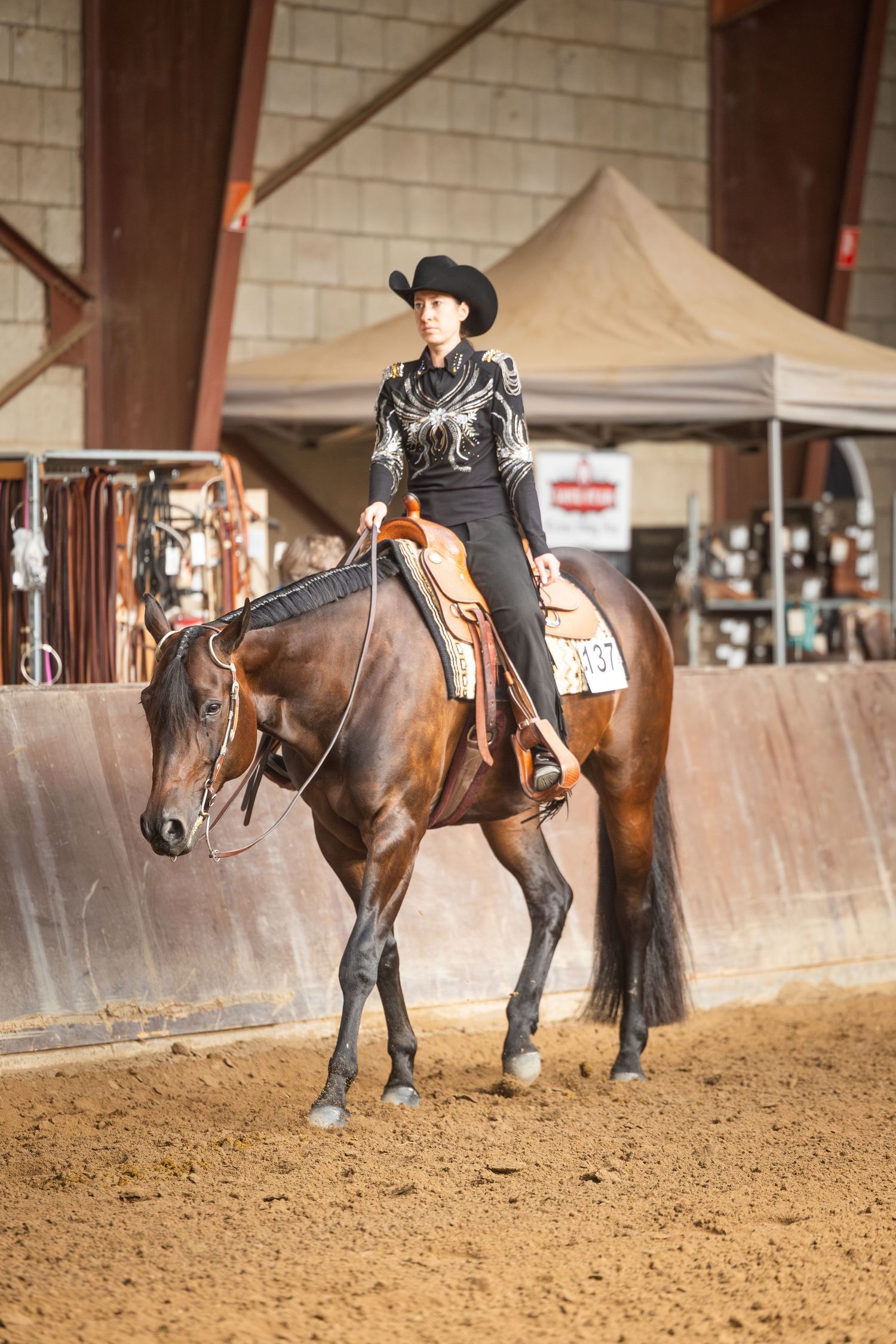 Woman in black cowboy hat and western attire riding a bay horse in an indoor equestrian arena competition.