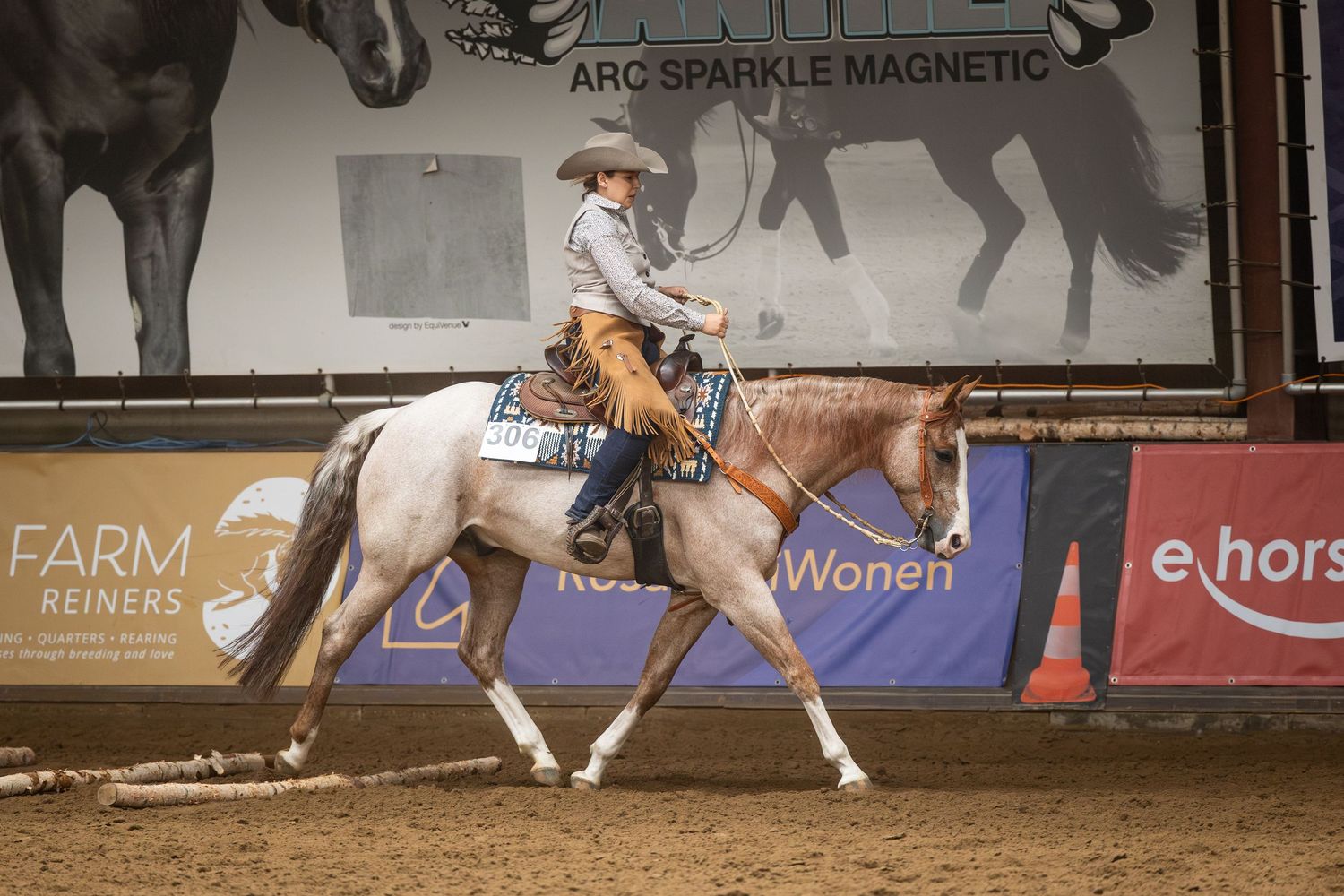 Rider on a roan horse competing in a reining event indoors, with sponsor banners visible in the background.