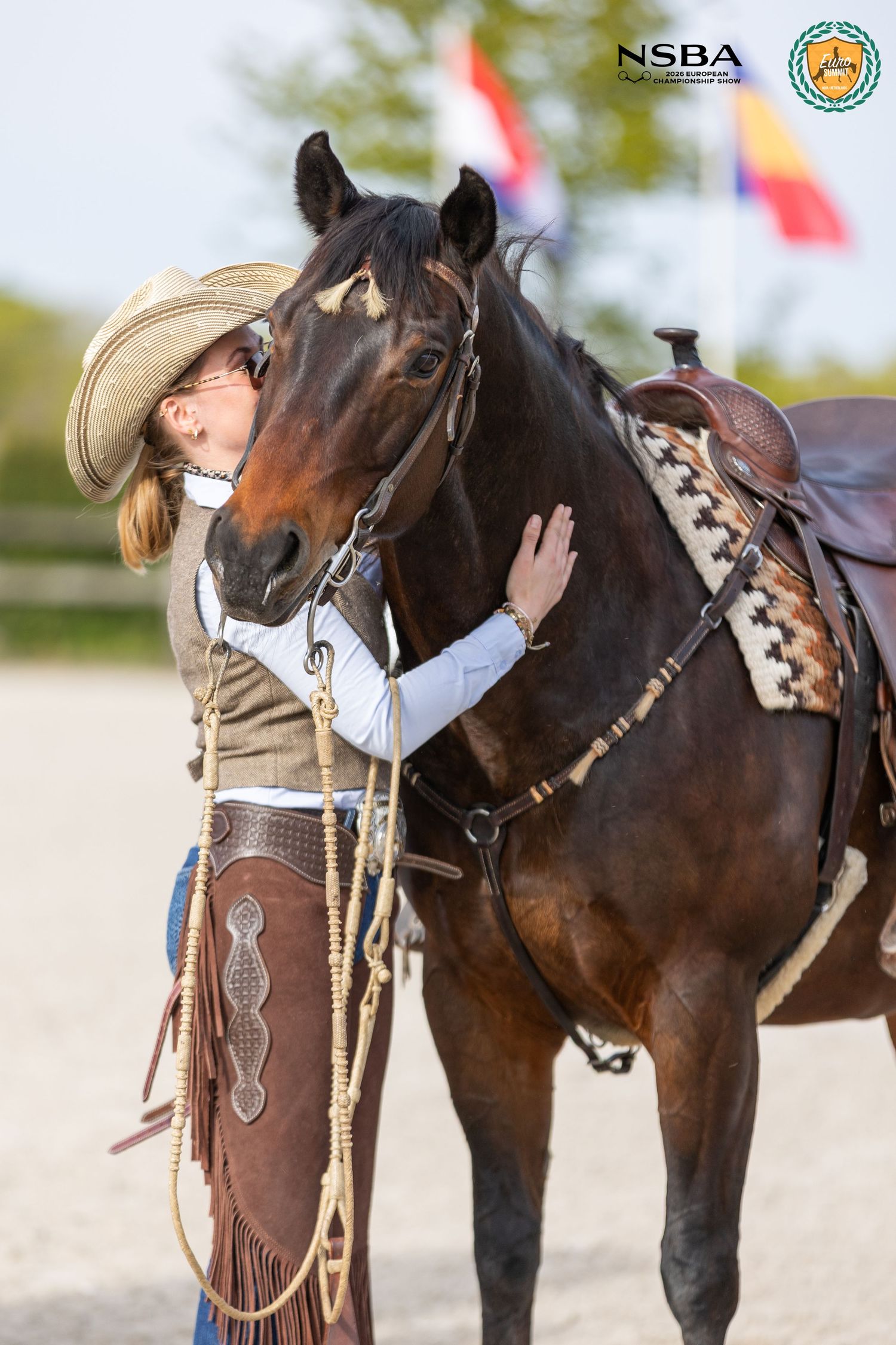 A cowgirl in a tan hat affectionately nuzzles a dark bay saddled horse outdoors.