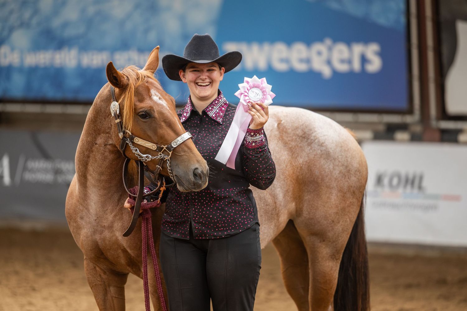 Woman in cowboy hat proudly holds ribbon rosette beside chestnut horse at indoor equestrian competition.