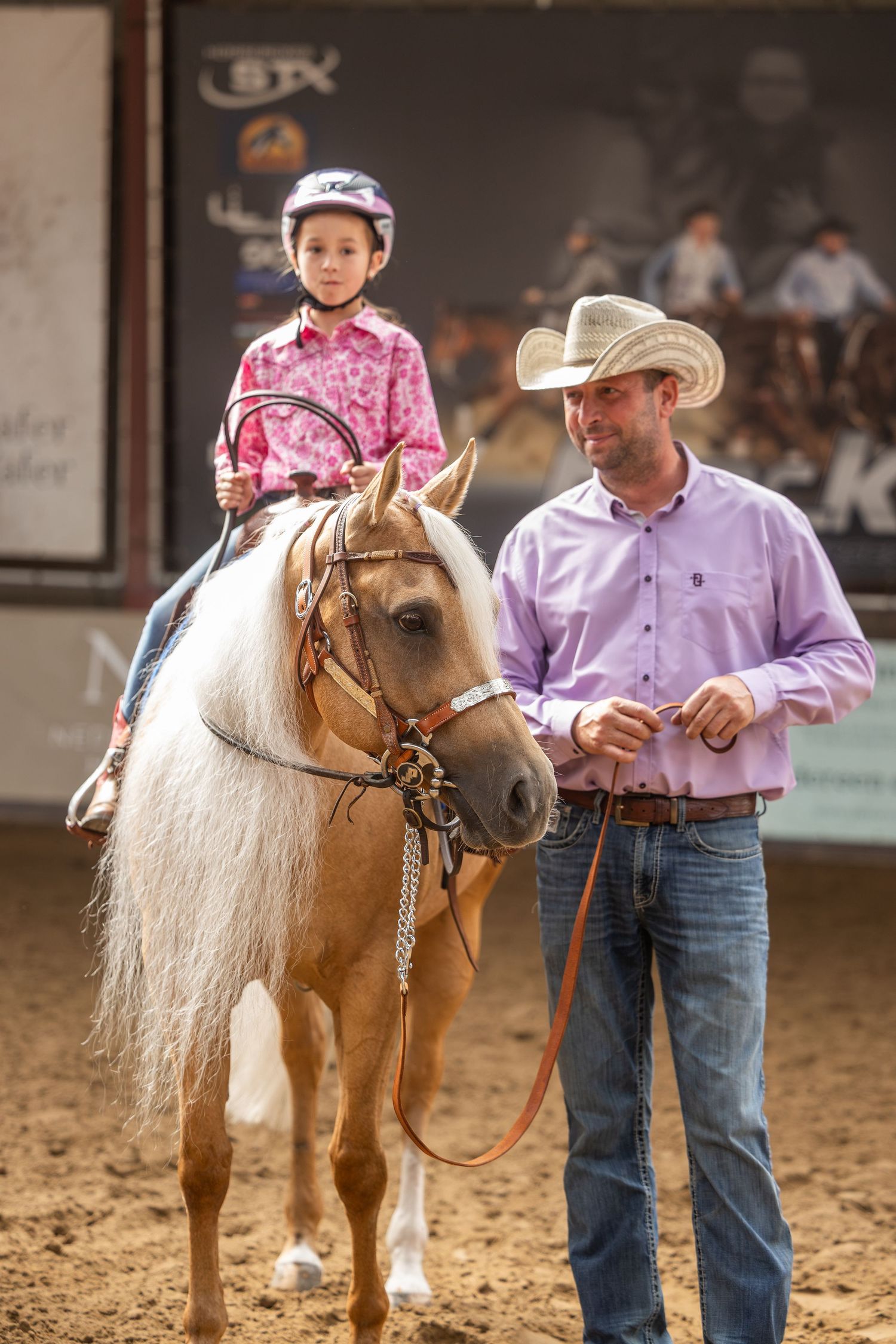 A young girl in a pink shirt and helmet rides a palomino pony led by a man in a cowboy hat at an equestrian event.