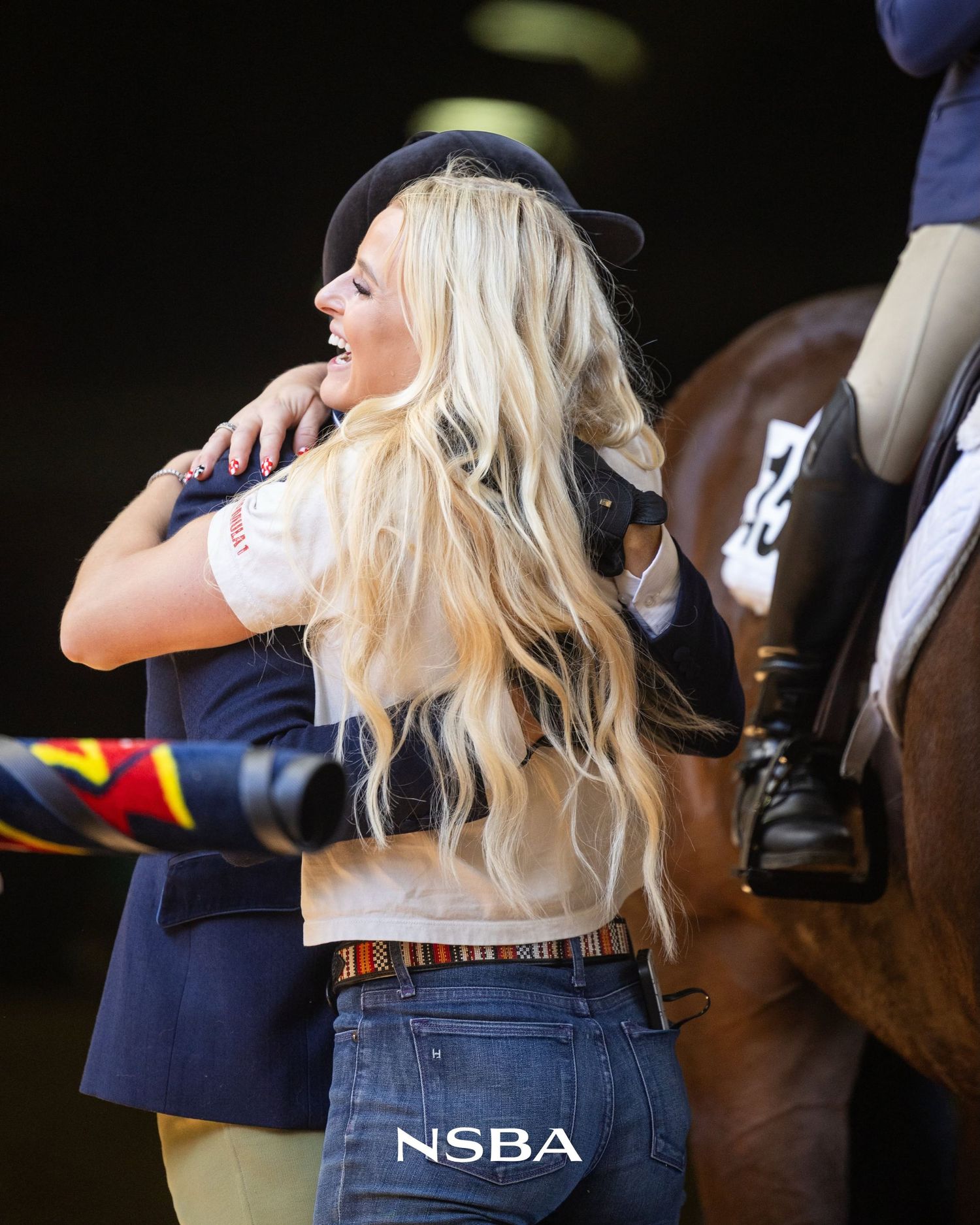Blonde woman in NSBA jeans hugging someone at an equestrian event near a brown horse.