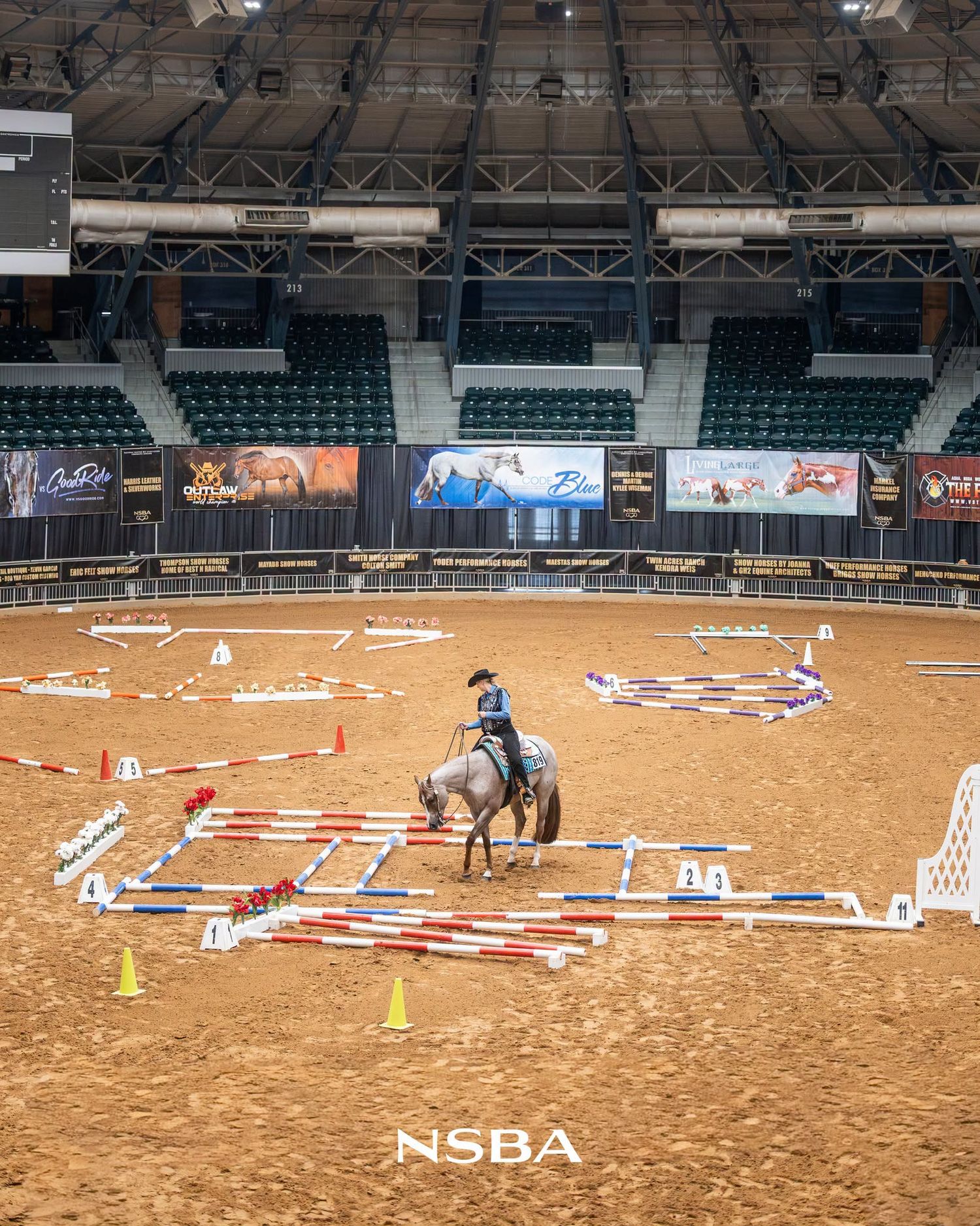 Young rider on a gray horse navigating a show jumping course in an indoor arena at an NSBA equestrian event.