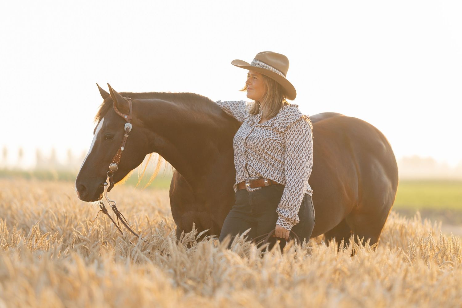 Woman in cowboy hat standing beside a dark bay horse in a golden wheat field at sunset.