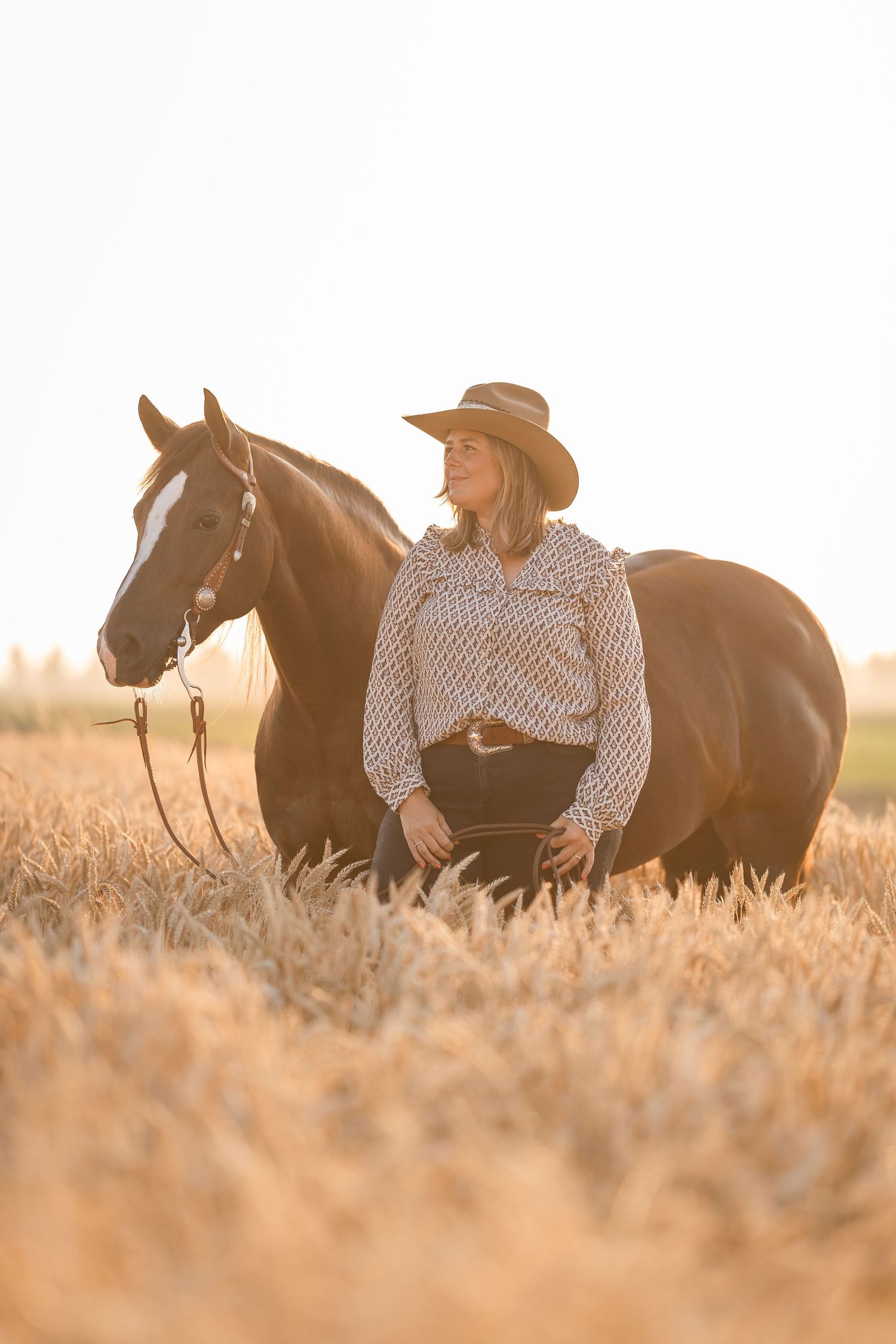 A woman in a cowboy hat and patterned shirt stands beside a dark horse in a golden wheat field at sunset.