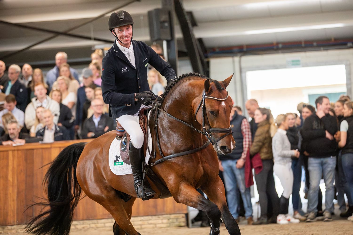 Professional equestrian riders compete in an indoor arena with brown horses while spectators watch from the sidelines.