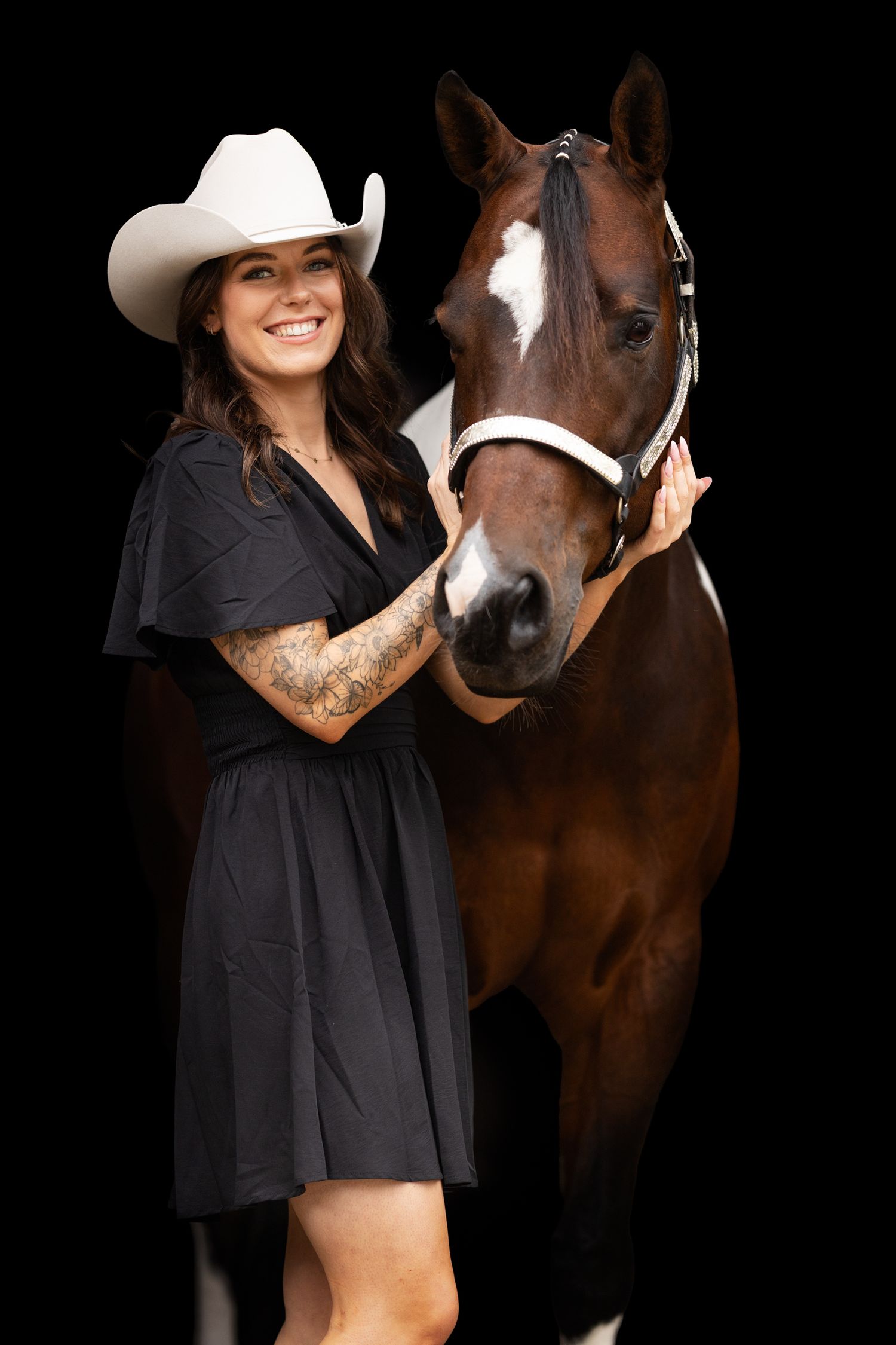 Woman in black dress and white cowboy hat stands beside brown horse in studio.