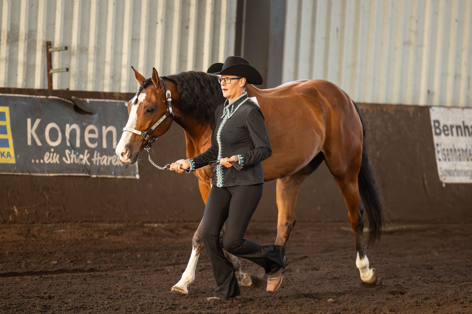 Handler leading a bay horse in western show attire during an indoor equestrian event.
