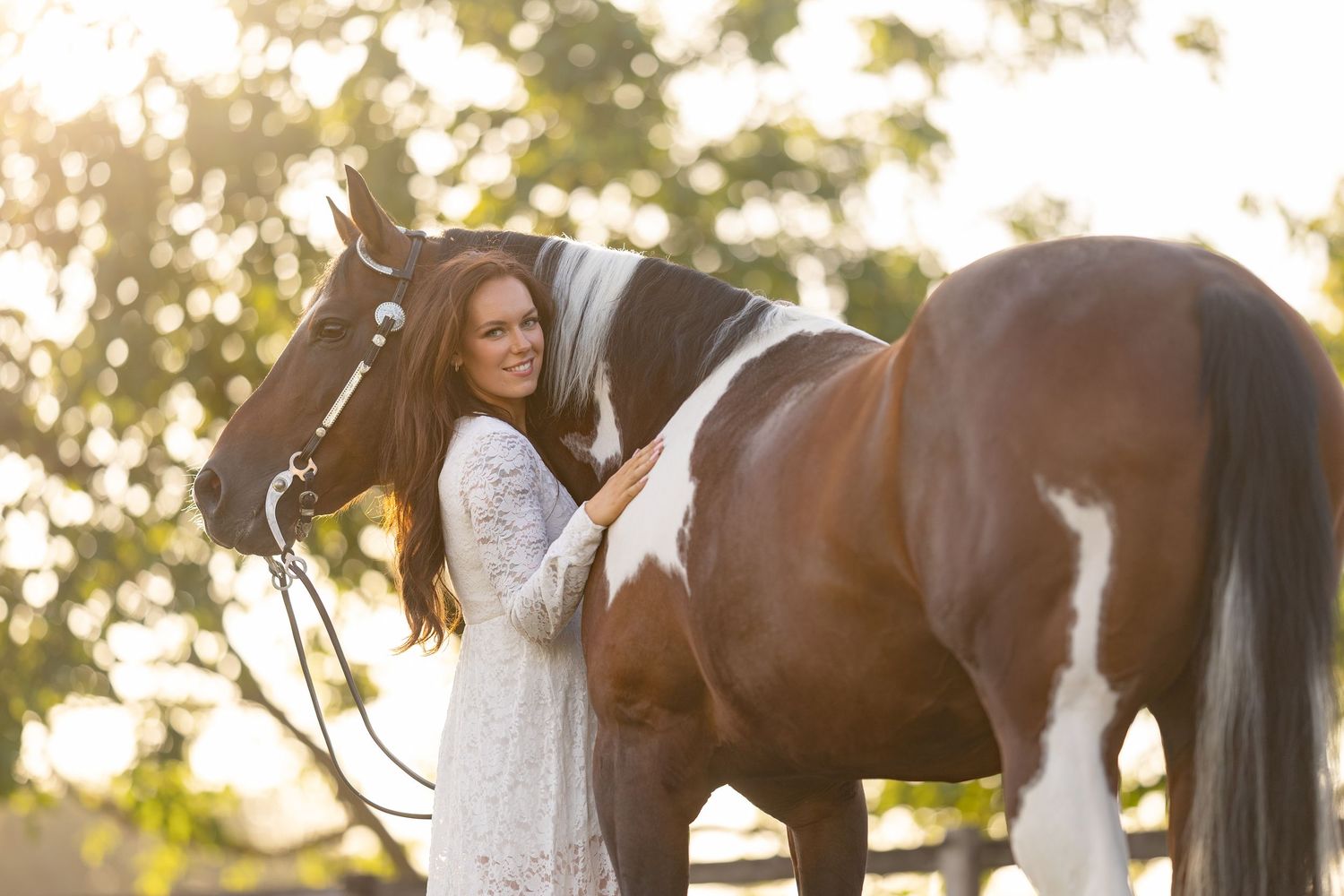Young woman in white lace dress smiling beside a brown and white paint horse outdoors in golden sunlight.