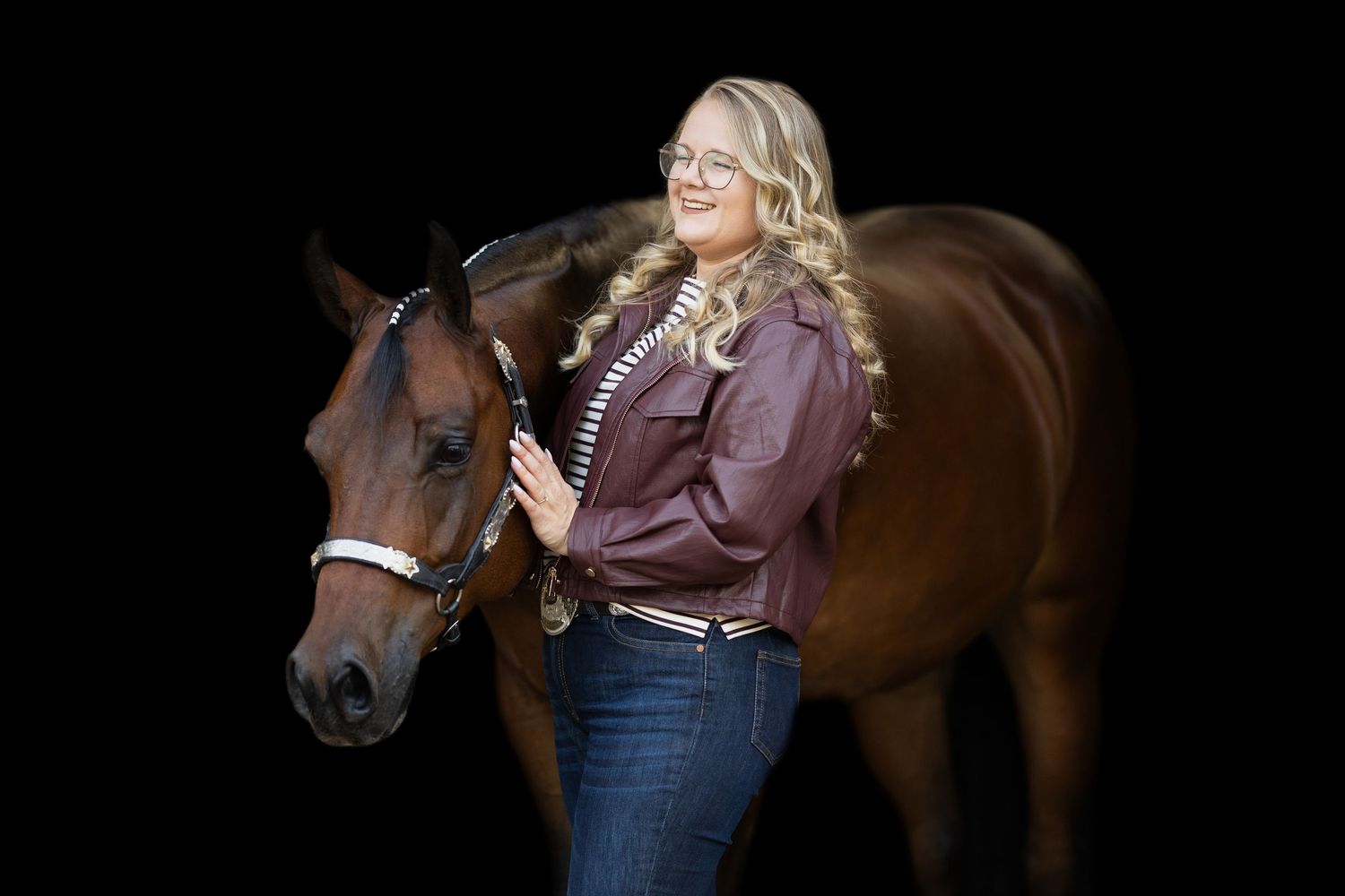 Blonde woman in maroon jacket and jeans posing with a chestnut horse against a black background.