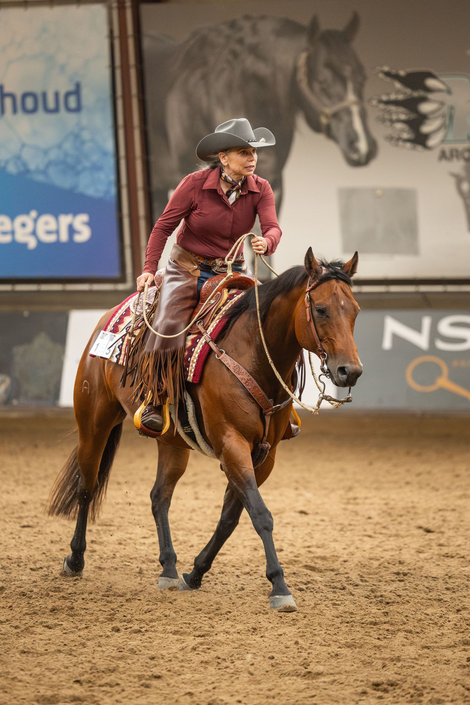 A woman in a cowboy hat rides a chestnut horse in an indoor equestrian arena during a western riding competition.