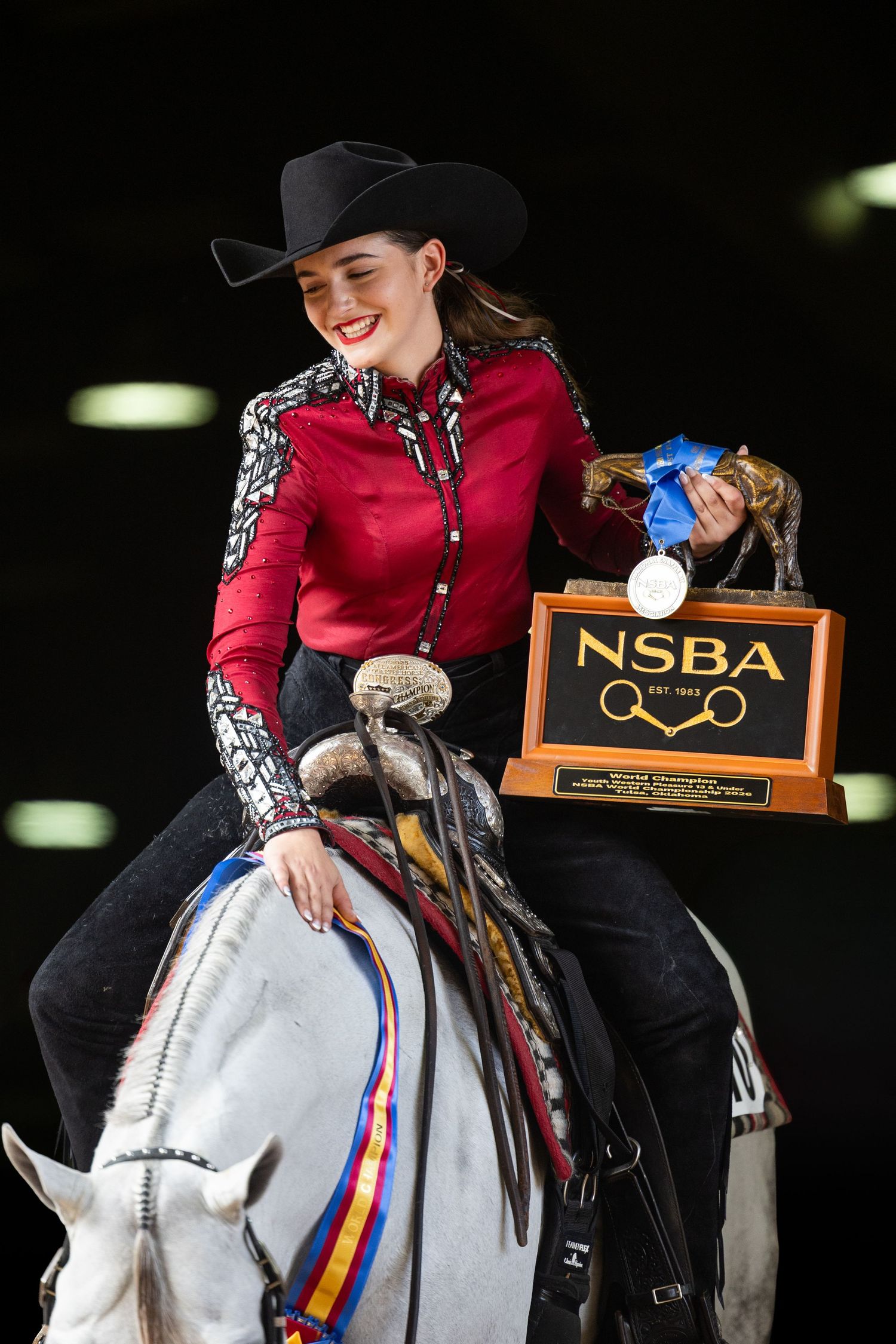 Young woman in red shirt and black cowboy hat on white horse holding NSBA trophy and blue ribbon.