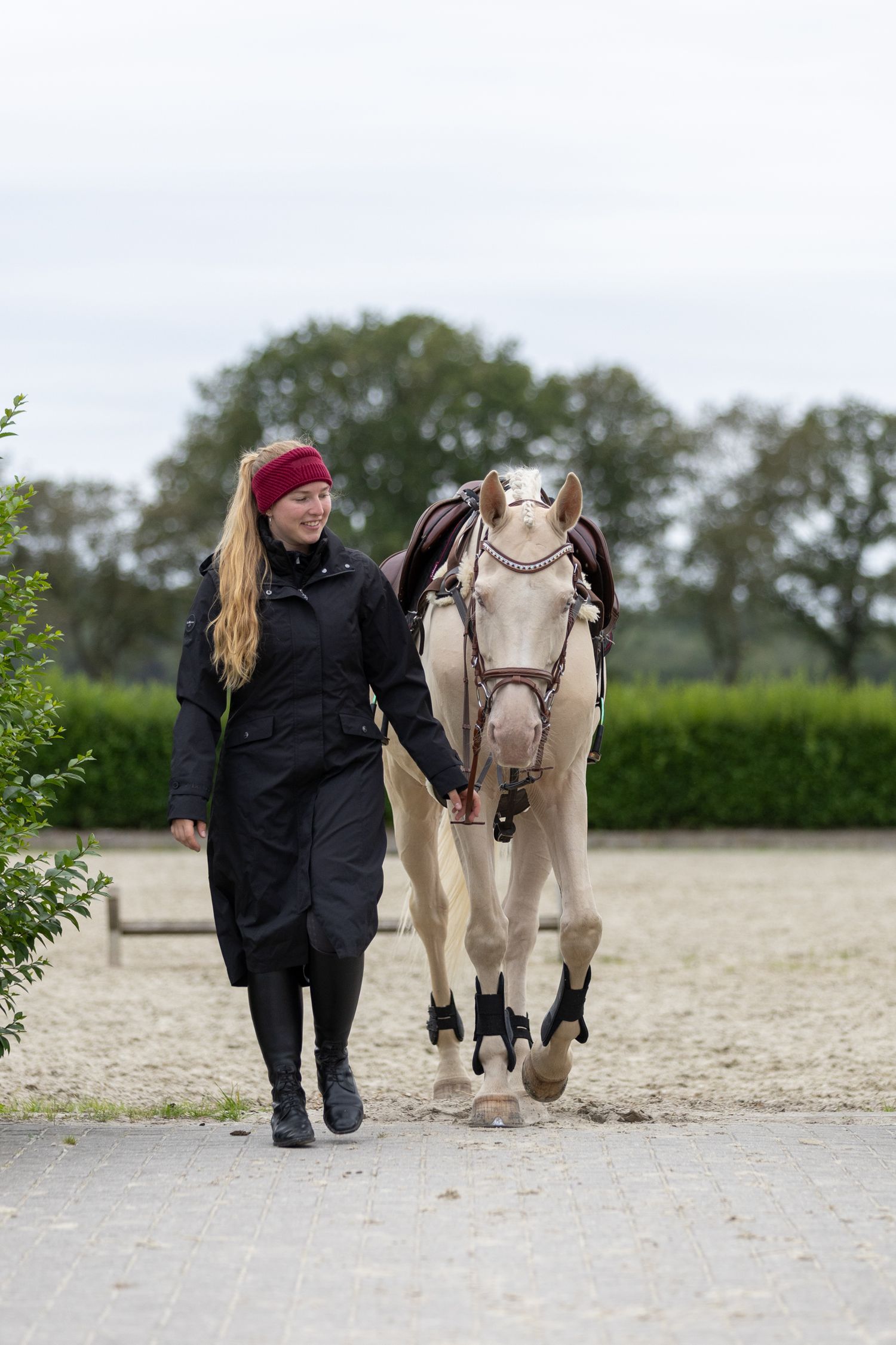 Person in black coat walking alongside a cream-colored pony on a sandy path.