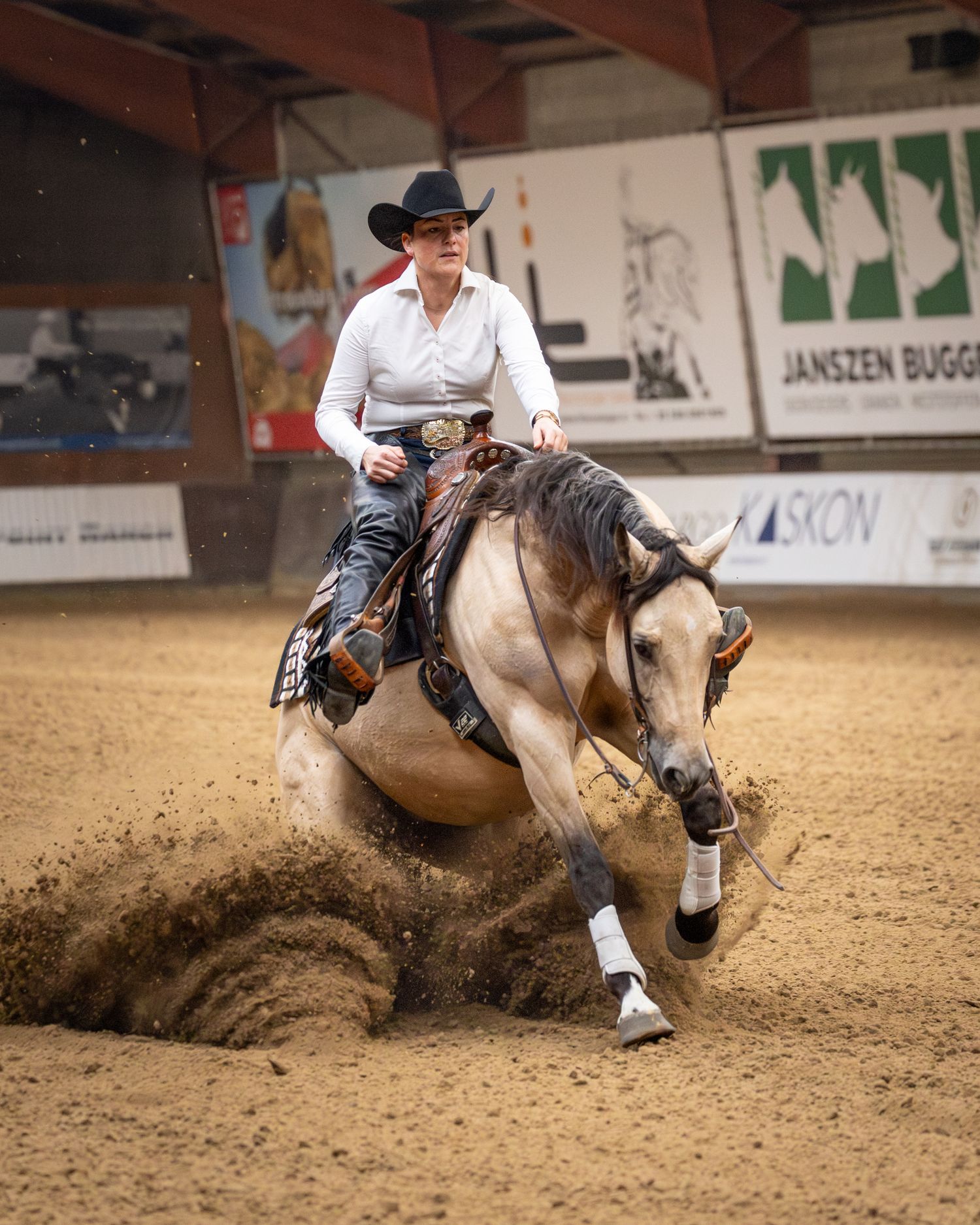 A western rider performs a sliding stop on a buckskin horse in a sandy arena, creating dramatic dust clouds.