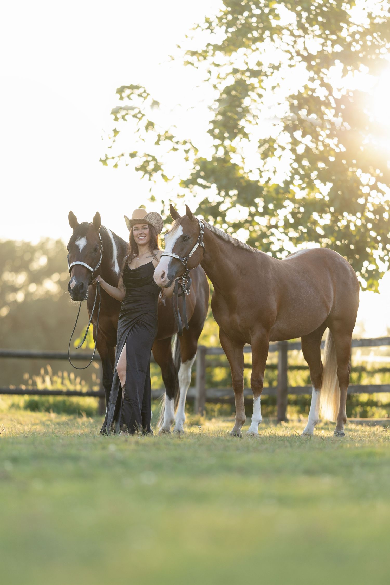 Woman in black dress and cowboy hat stands with two horses at golden hour on a sunny farm.