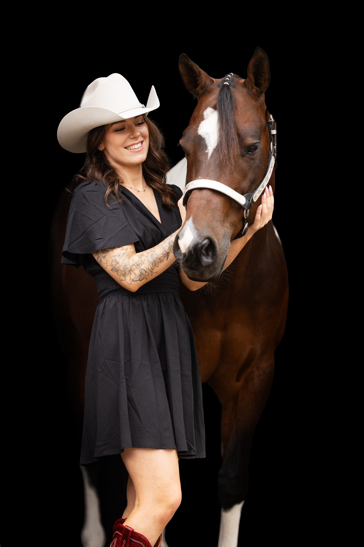 Woman in black dress and cowboy hat poses with brown and white horse against dark background.