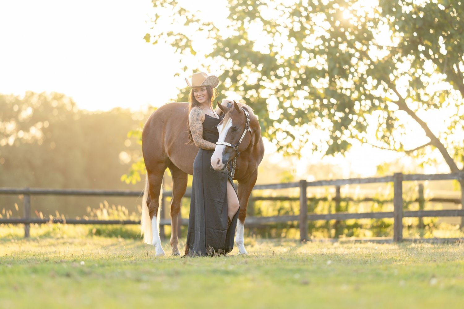 Woman in black dress and cowboy hat stands with chestnut horse in sunlit pasture with wooden fence.