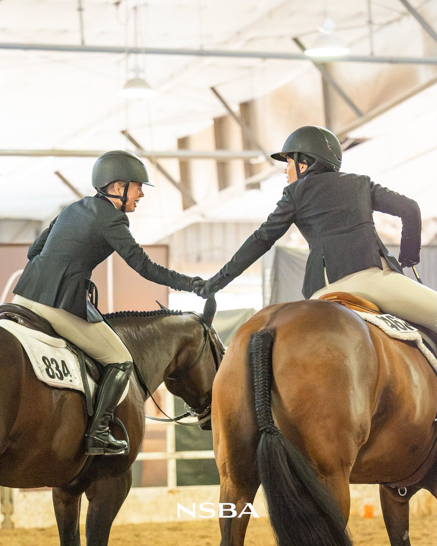 Two equestrians in black jackets shake hands atop bay horses in an NSBA indoor arena.
