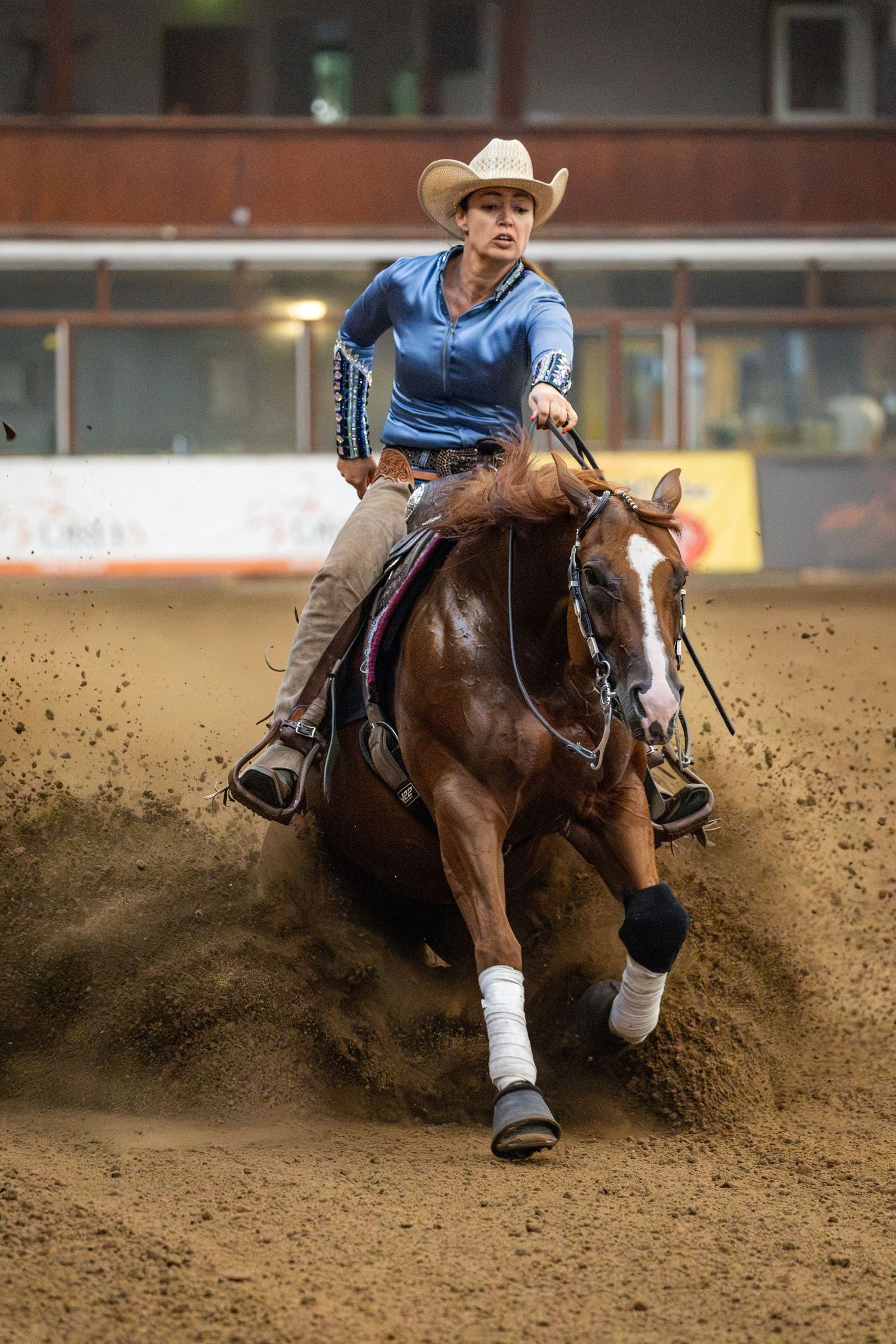 Cowgirl in blue shirt and white hat riding a chestnut horse at full gallop in an indoor rodeo arena.