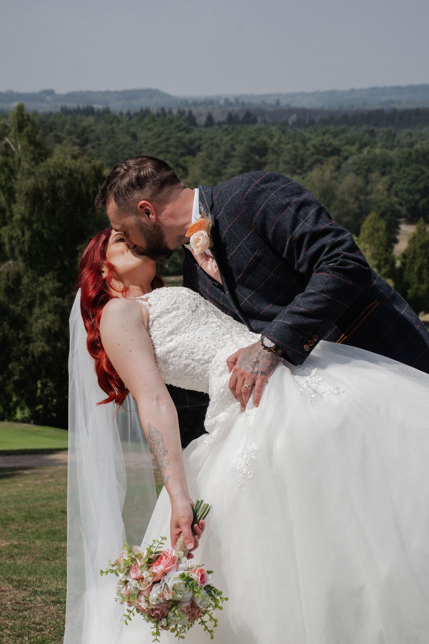 Groom dips red-haired bride for a kiss outdoors with scenic forest landscape behind them.