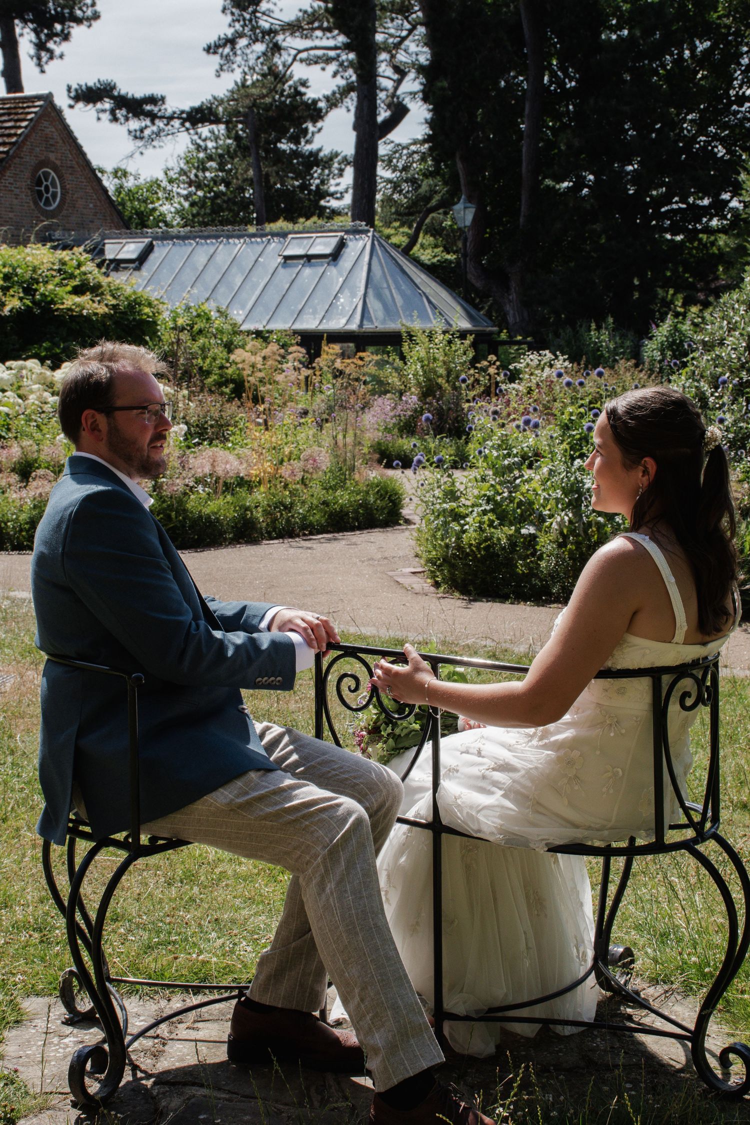 A bride and groom sit facing each other on decorative iron chairs in a lush garden during their wedding day.