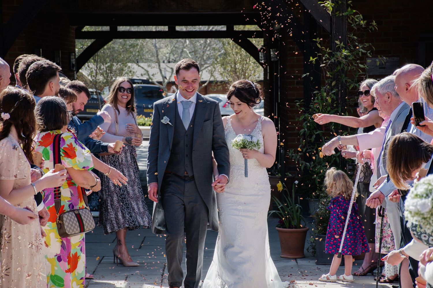 A newlywed couple walks through a confetti shower surrounded by cheering guests at their wedding. Cottesmore Golf and Hotel