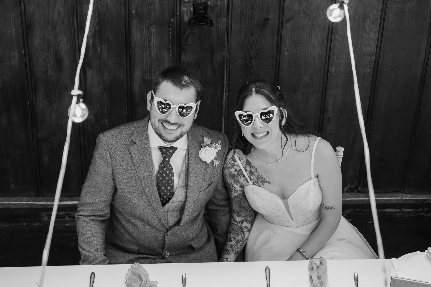 Black and white photo of smiling bride and groom wearing heart-shaped sunglasses at their wedding reception.