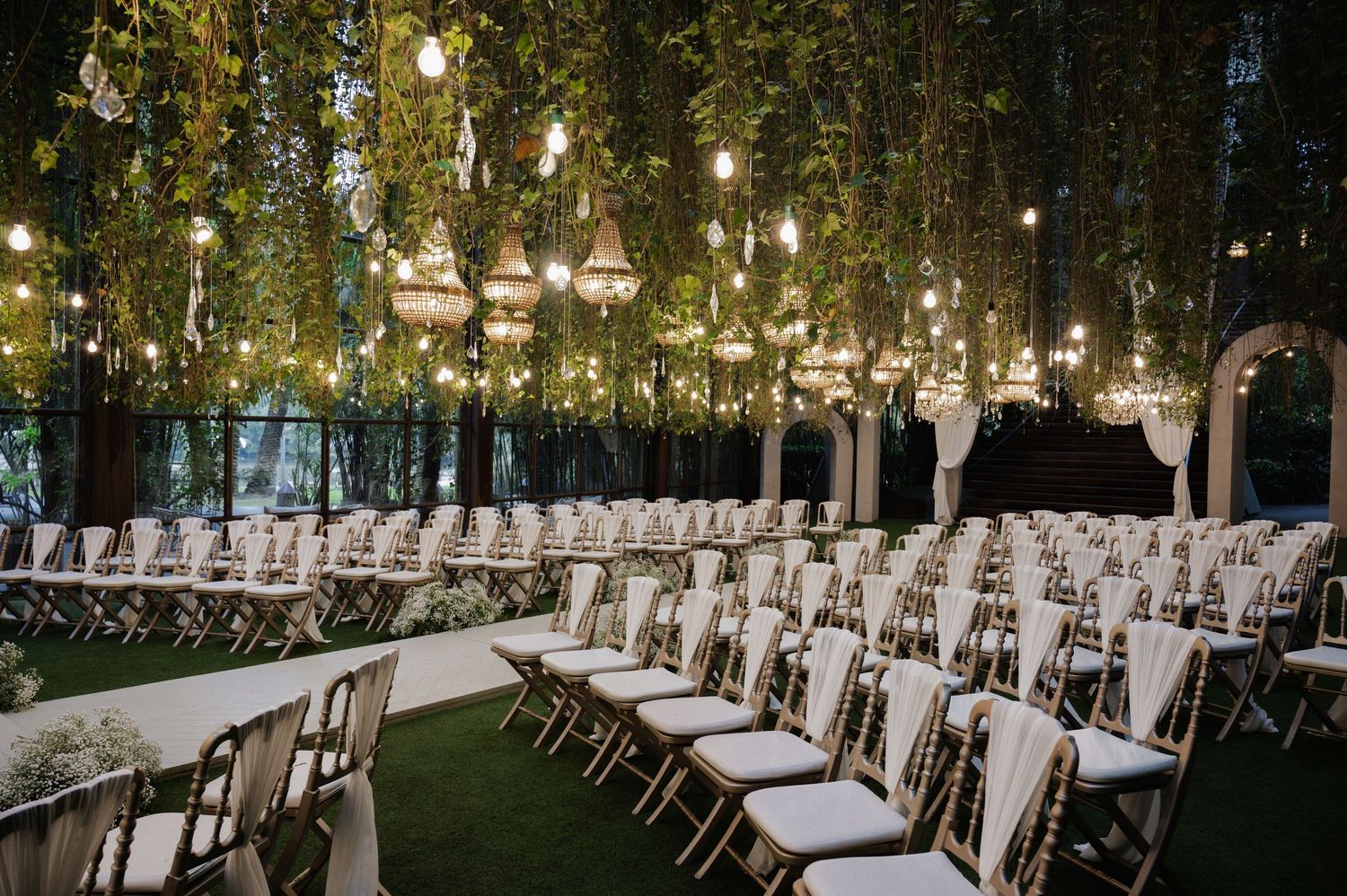 Elegant outdoor wedding ceremony setup with string lights, hanging lanterns, lush greenery canopy, and rows of white chiavari chairs.