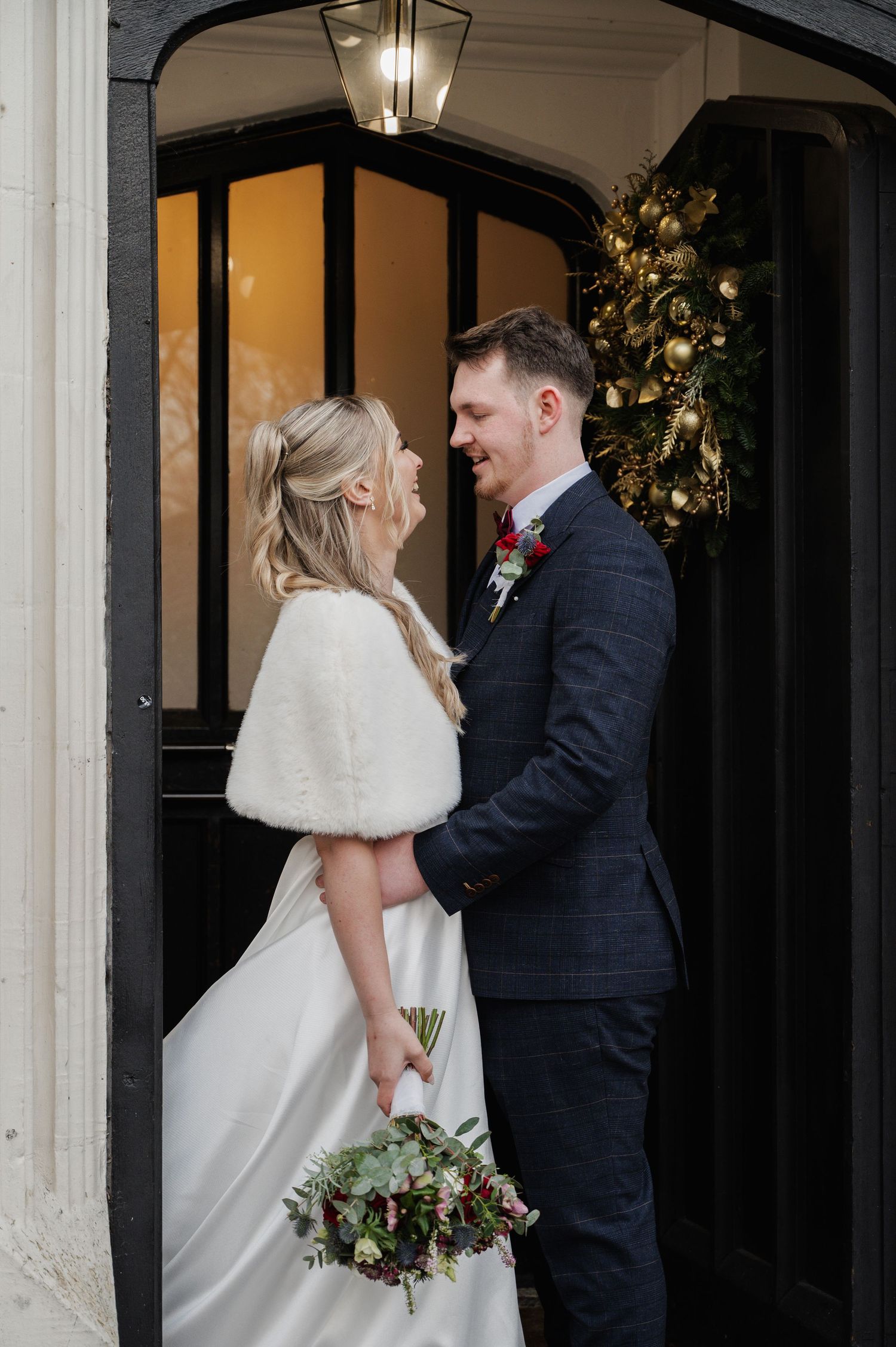 Bride in white fur stole and groom in navy suit laughing together at Christmas decorated doorway.