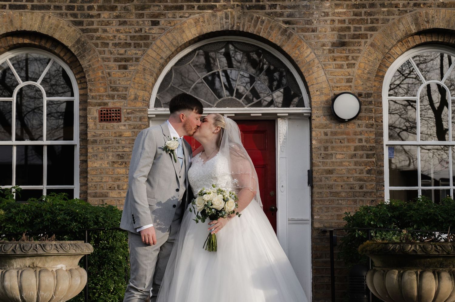 A bride and groom share a romantic kiss outside a brick building with arched windows on their wedding day.