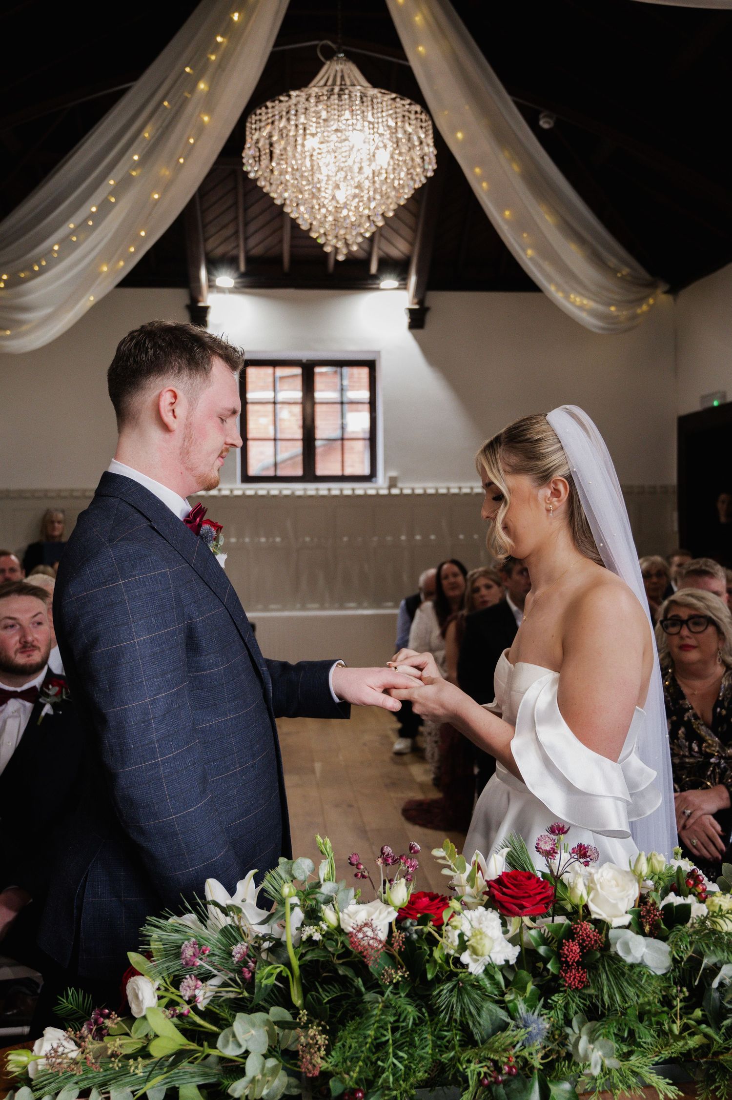 Bride places wedding ring on groom's finger during ceremony beneath crystal chandelier with guests.