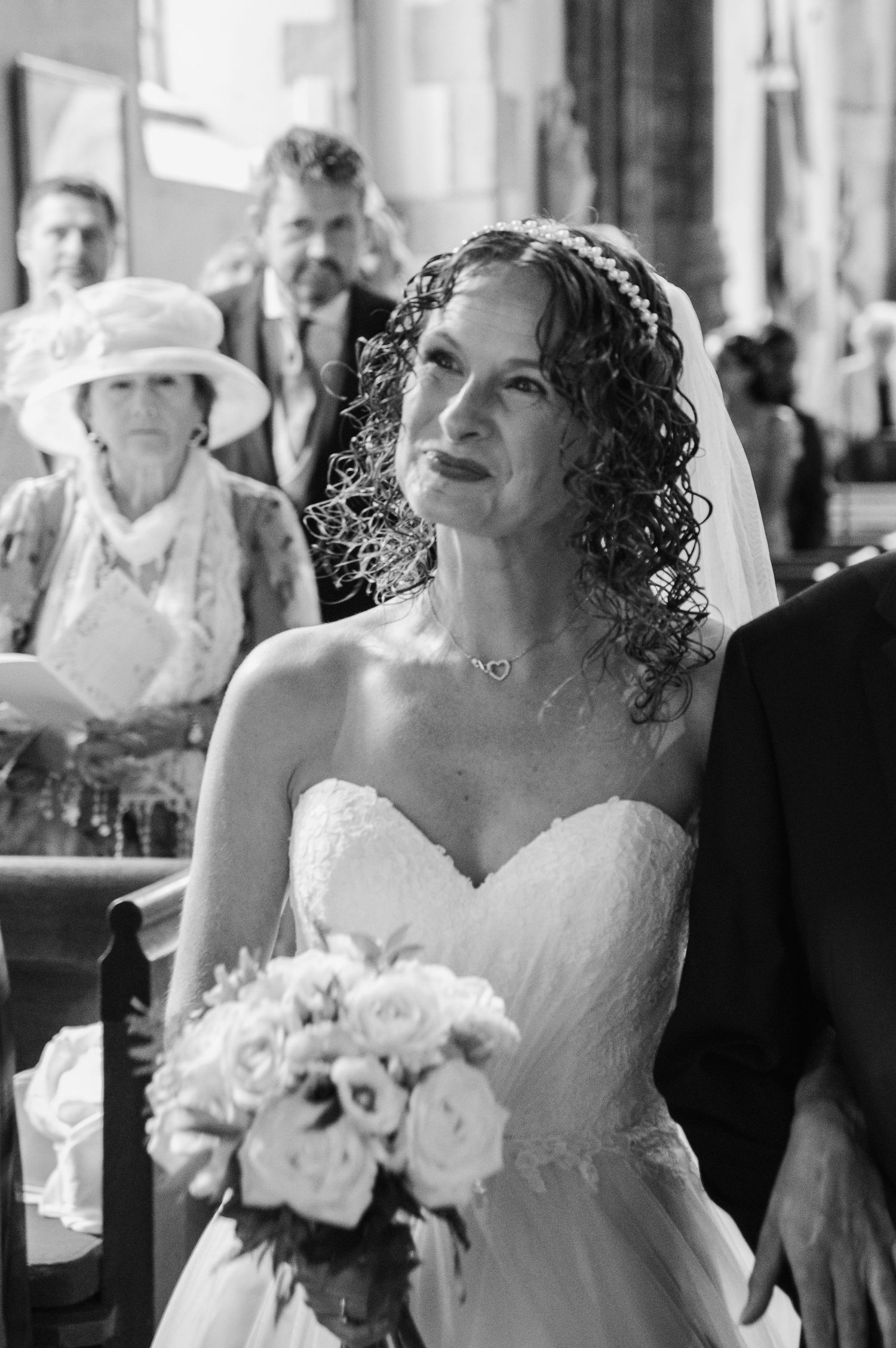 Elegant bride with curly hair holds white bouquet while walking down church aisle in black and white.