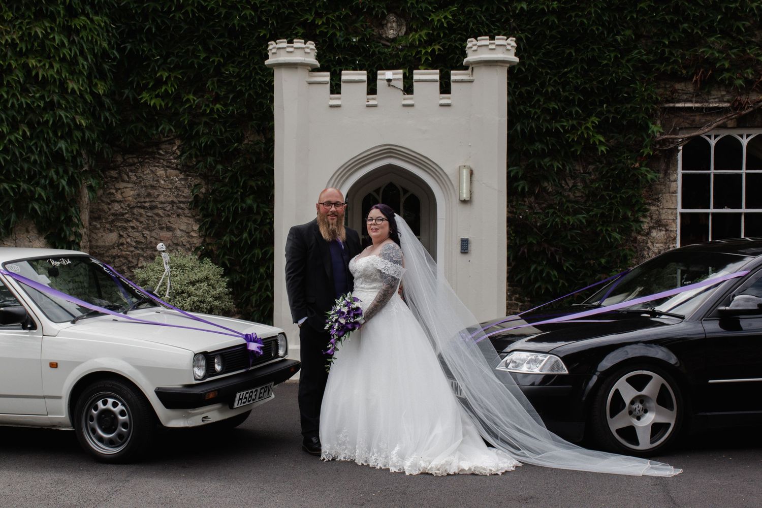 Bride in ball gown with purple bouquet and groom posing between two wedding cars at church.