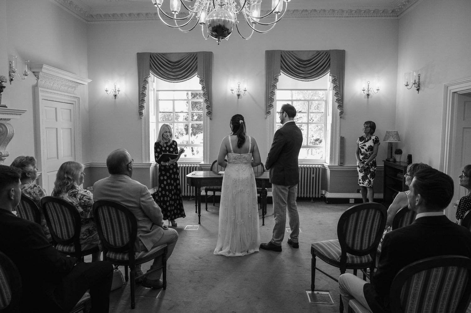A wedding ceremony in an elegant room with chandelier, guests seated as couple stands before an officiant.