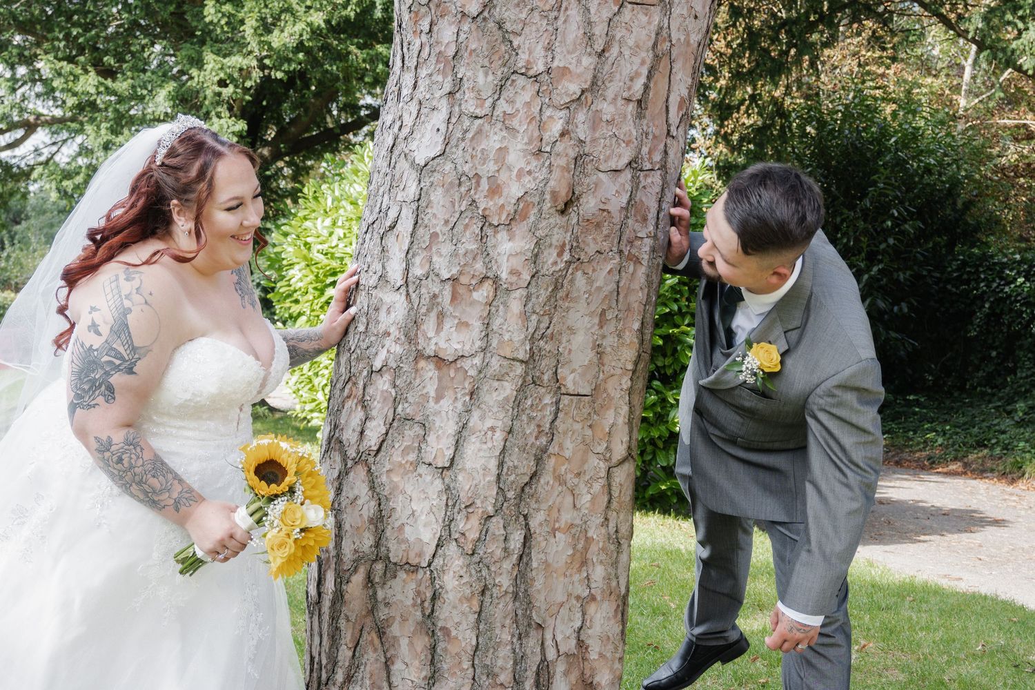 A tattooed bride holding sunflowers playfully peeks around a tree at her smiling groom outdoors.