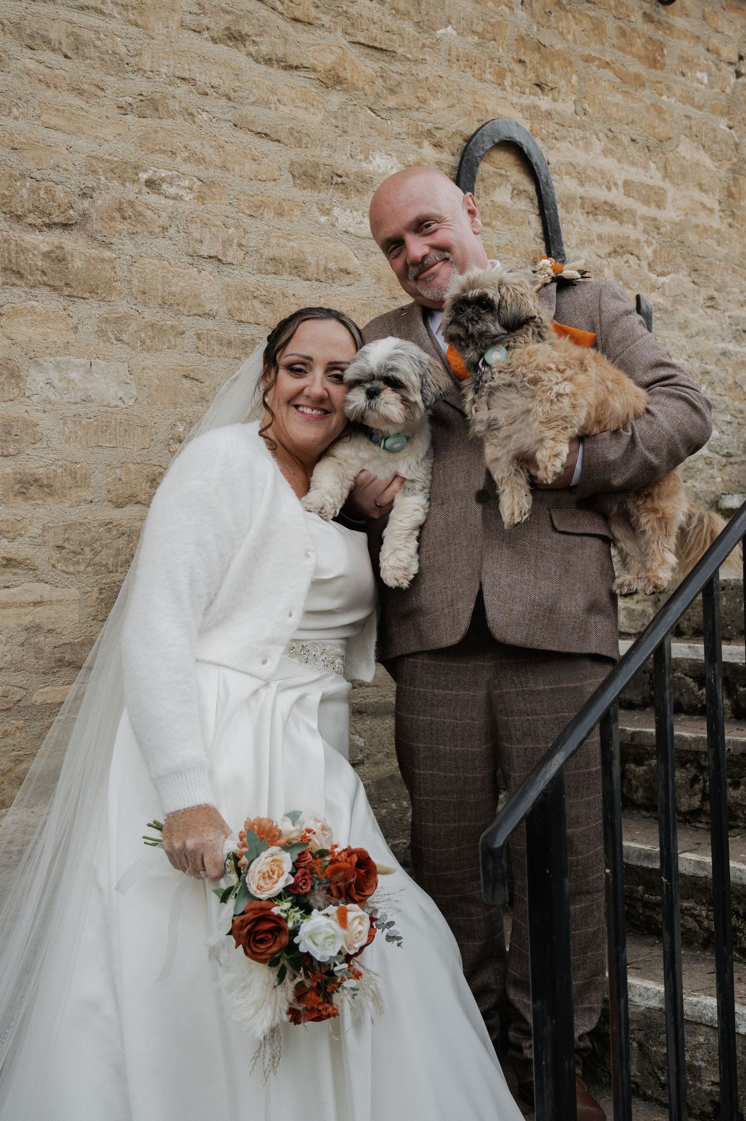 Happy bride and groom holding two small fluffy dogs on stone steps with a rustic stone wall backdrop.