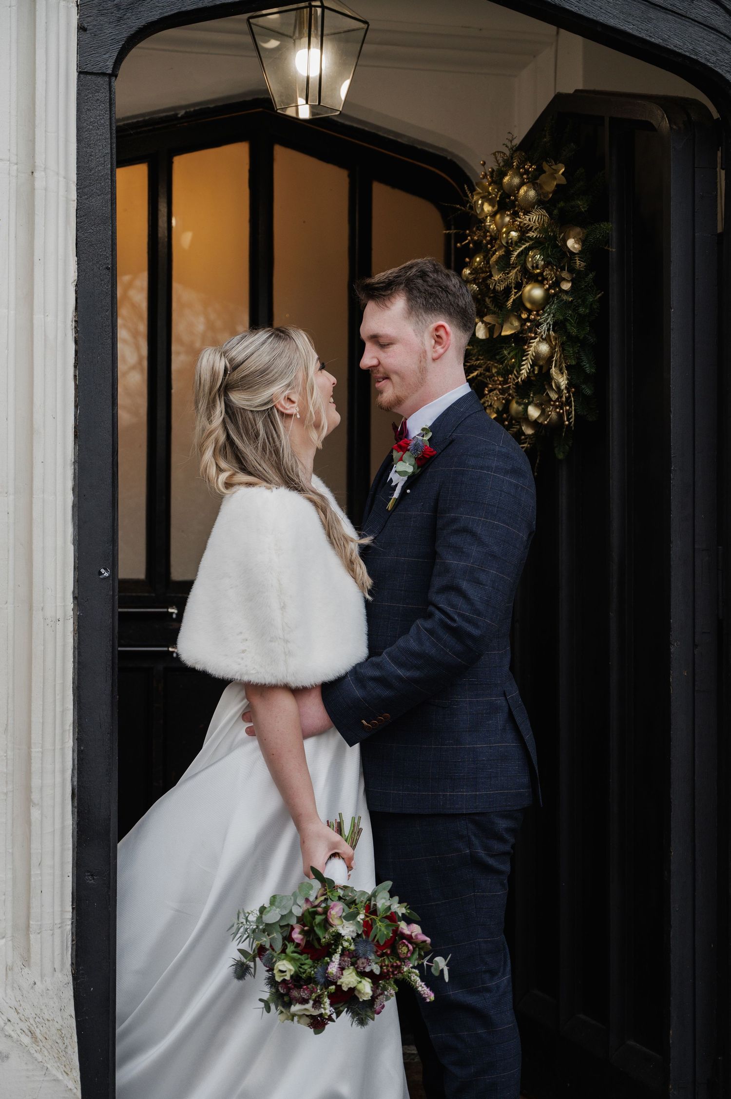 Couple embraces in doorway adorned with Christmas wreath, bride holding winter bouquet of dark florals.