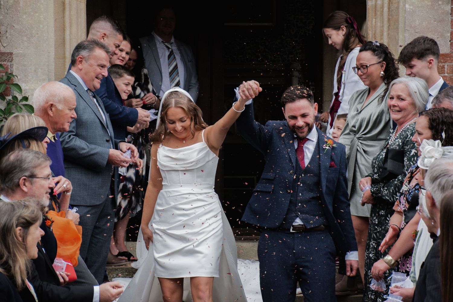 Joyful newlyweds walking through confetti shower with hands raised as wedding guests celebrate outside the venue.