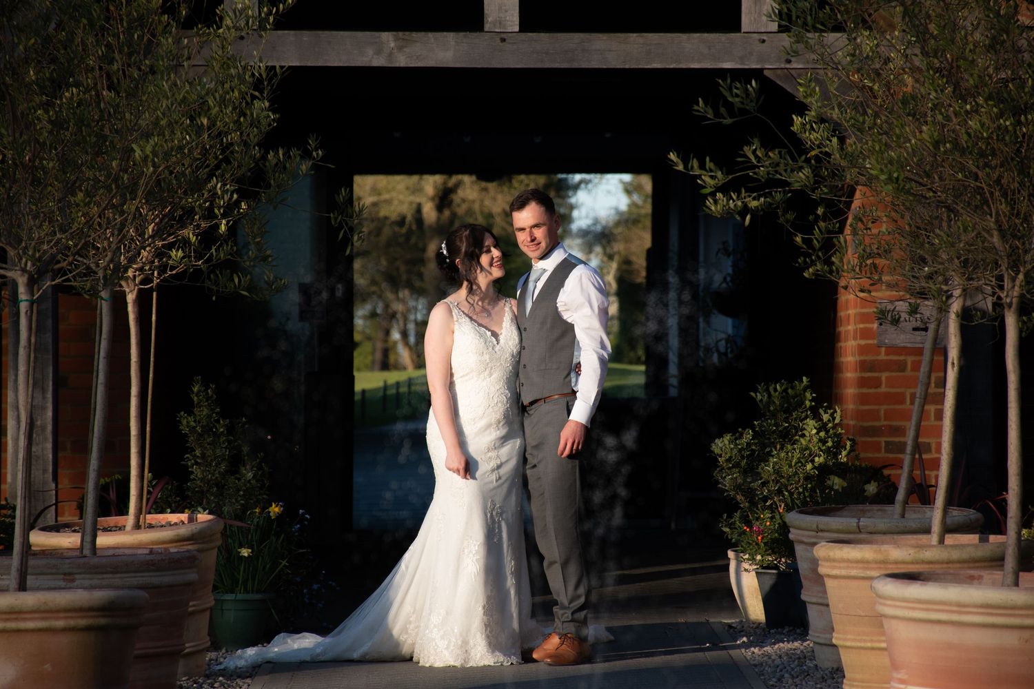 Bride and groom posing elegantly beneath a wooden archway flanked by potted olive trees at their wedding venue.