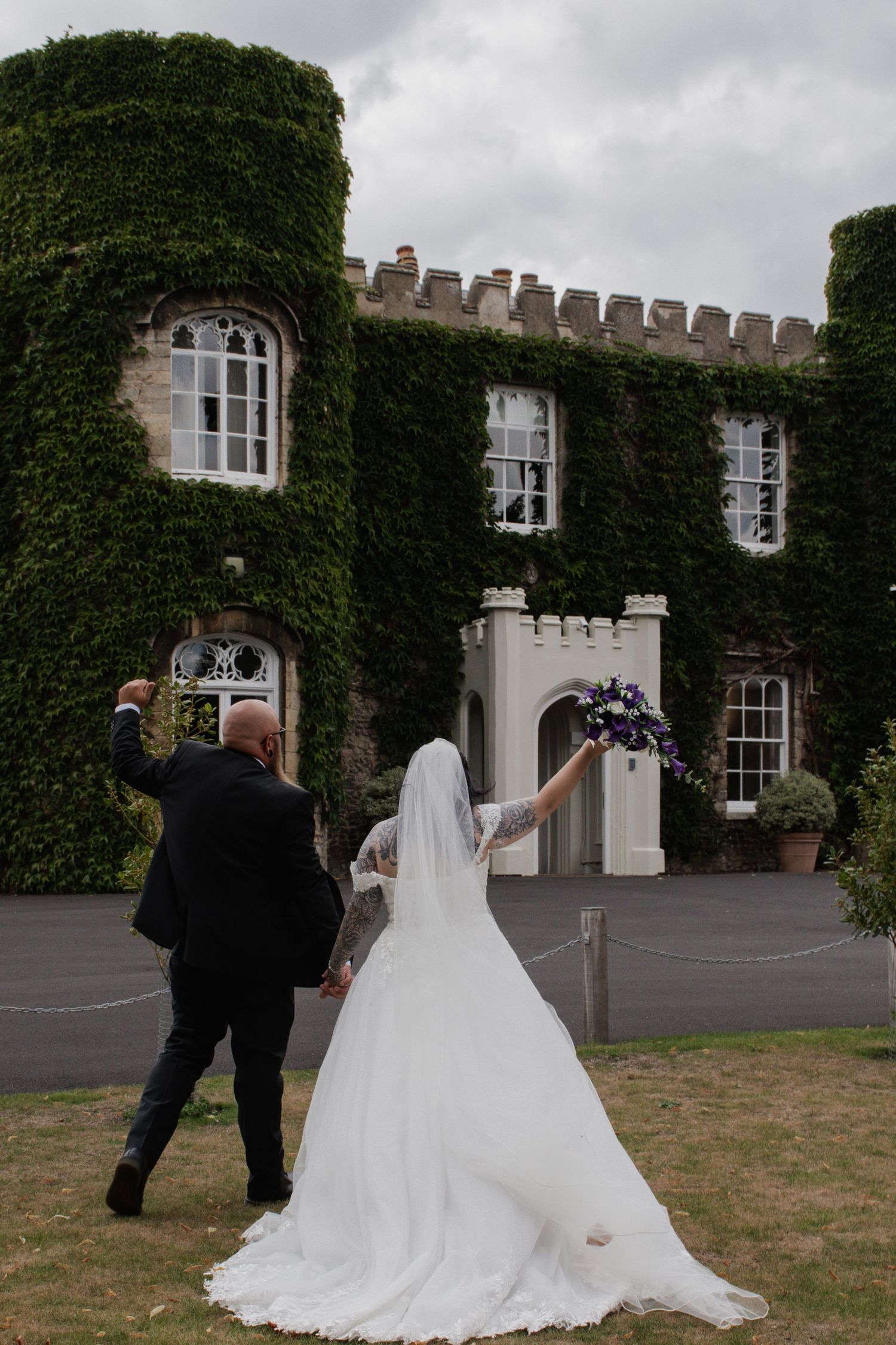 Bride and groom celebrating outside stunning ivy-covered castle wedding venue holding bouquet aloft.